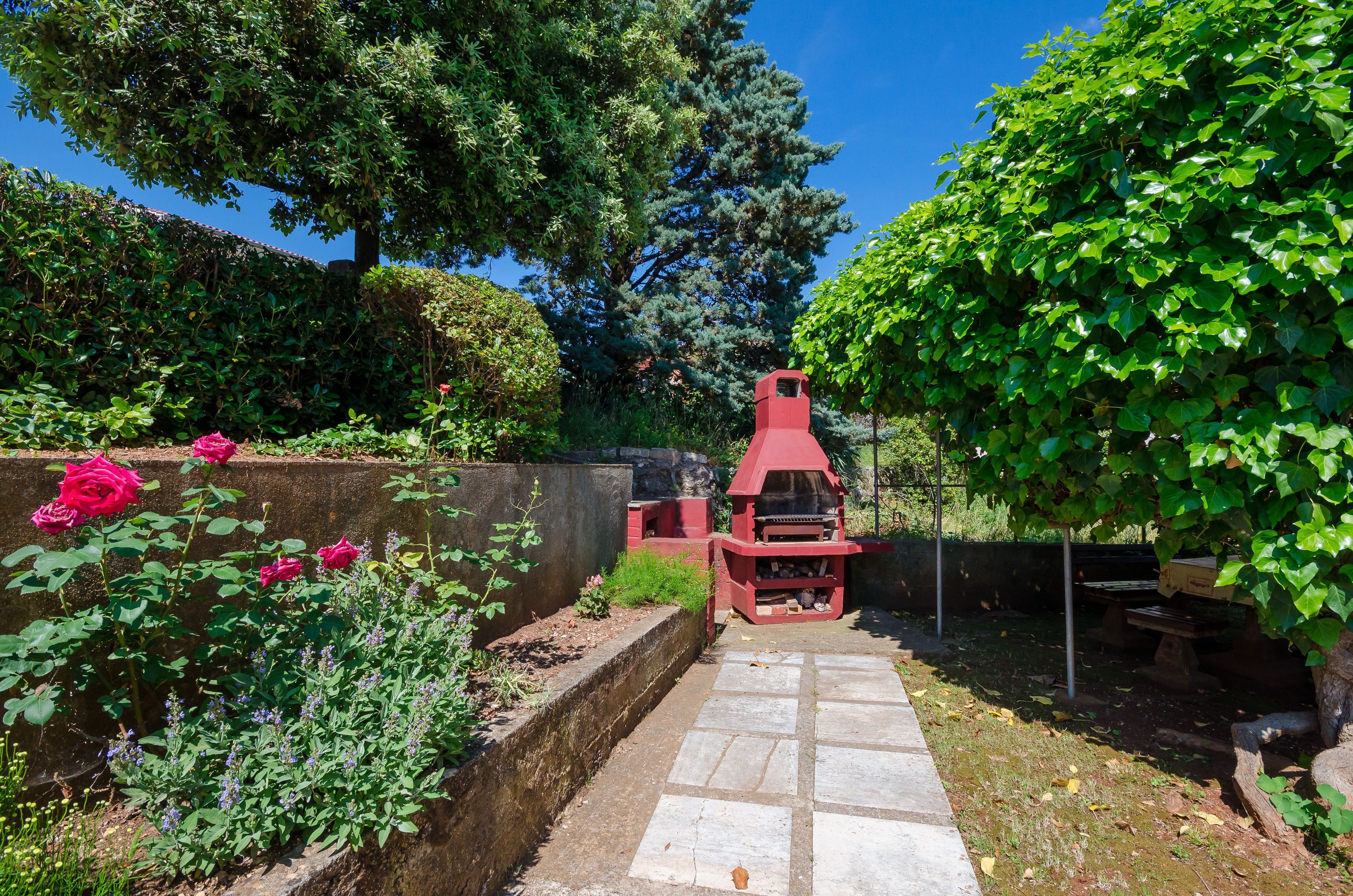 A garden with a stone pathway leading to a red barbecue area, surrounded by lush greenery and flowering plants.