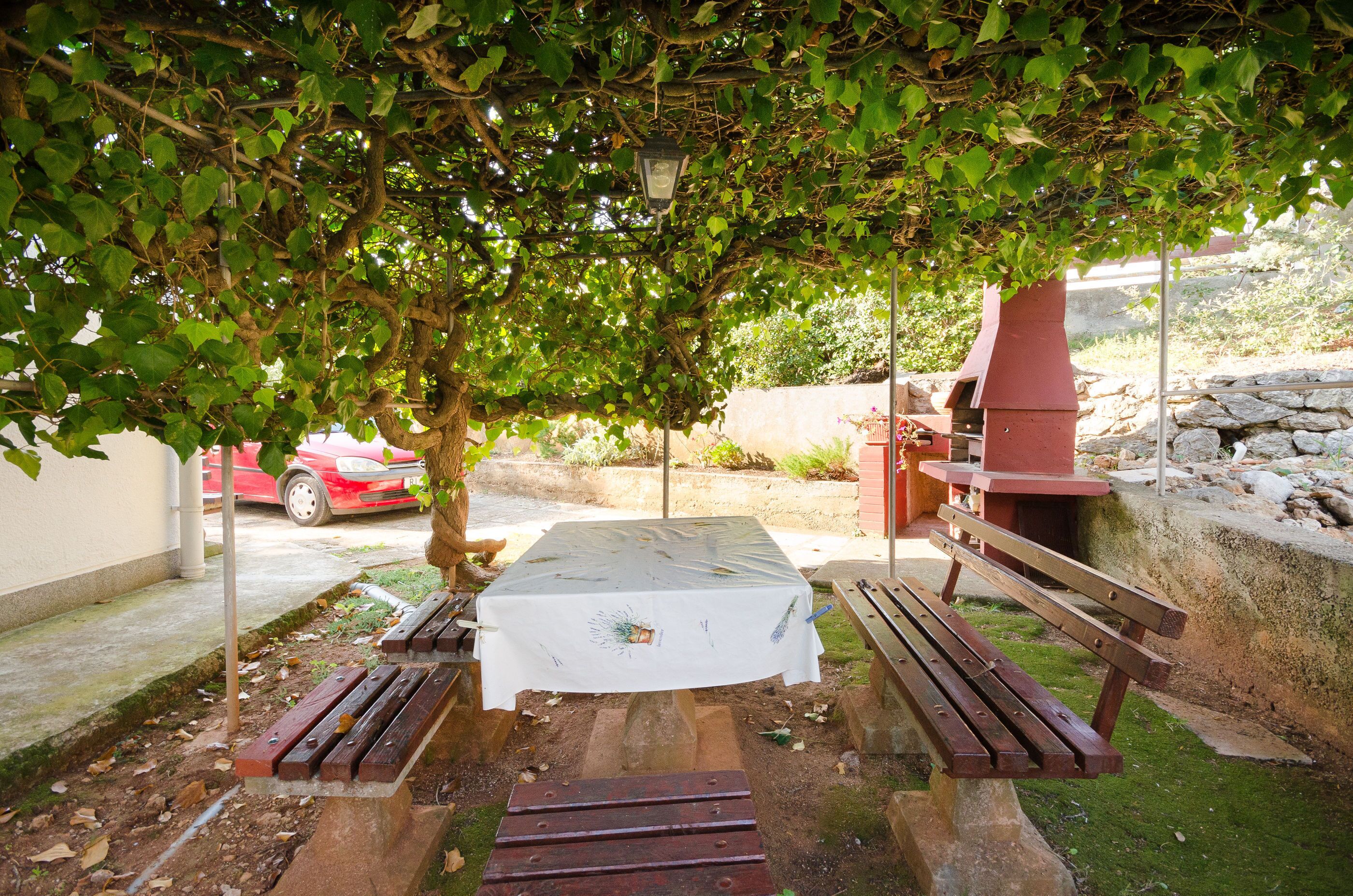 An outdoor dining area with a table and benches under a tree, a barbecue grill, and a red car in the background.