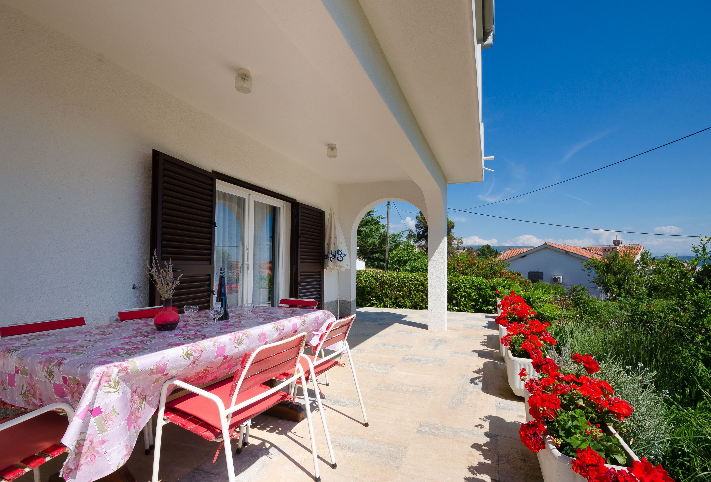 An outdoor patio with a table and chairs, a covered area with a view of the surrounding landscape, and a row of potted plants with red flowers.