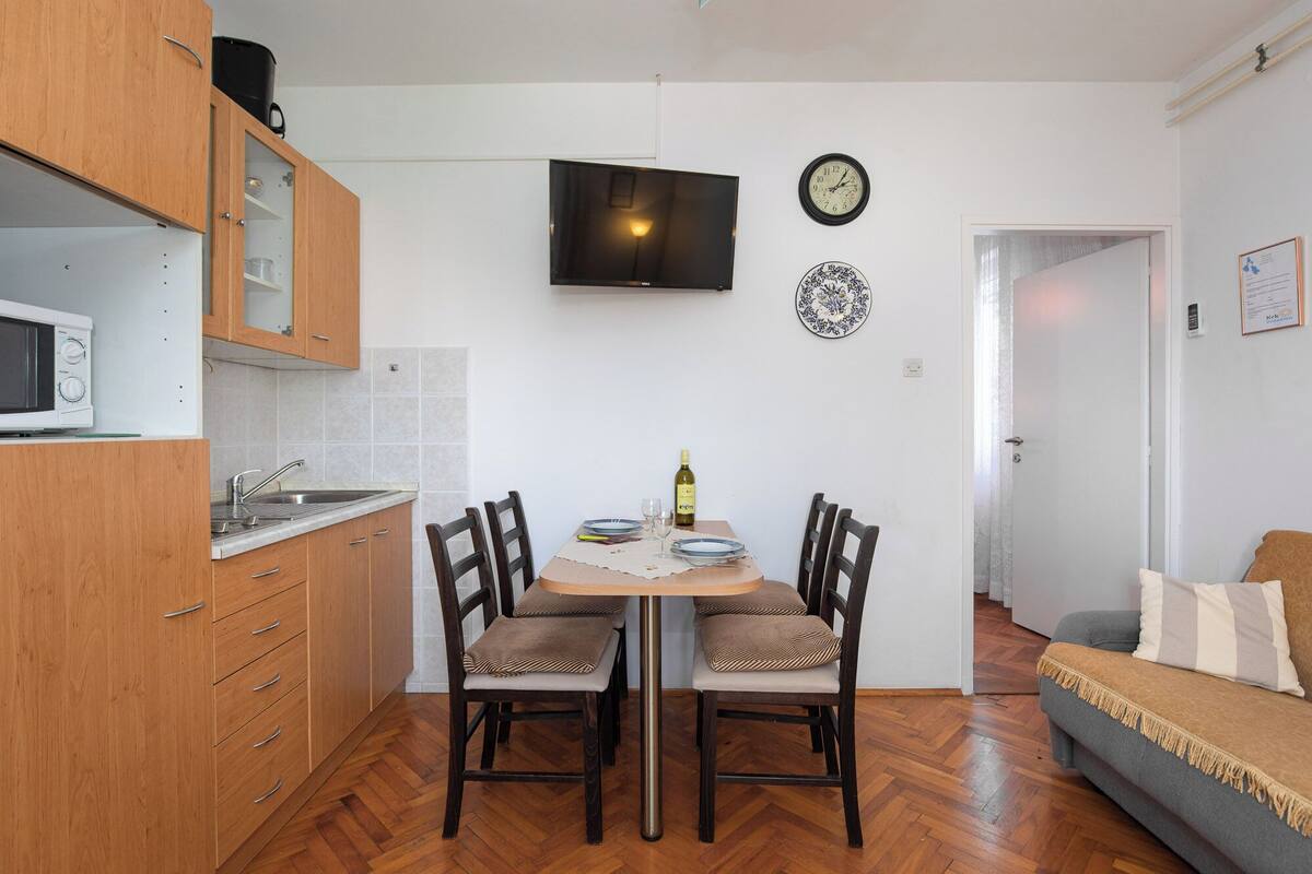 A compact kitchen with wooden cabinets, a microwave, and a dining table with chairs. A flat-screen TV is mounted on the wall above the table.