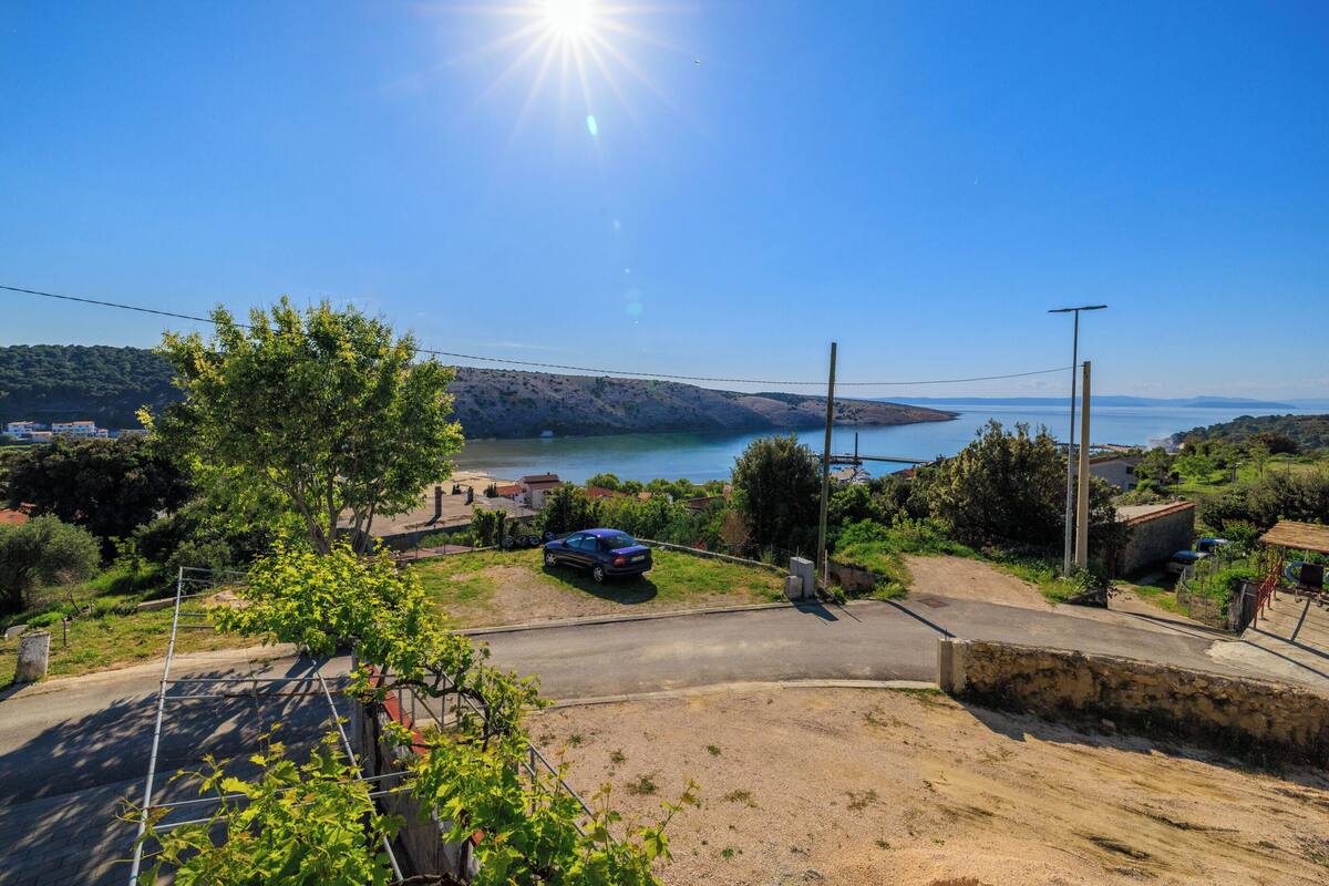 Une route côtière bordée d’une voiture garée, un ciel dégagé et une vue lointaine sur la mer et les collines.