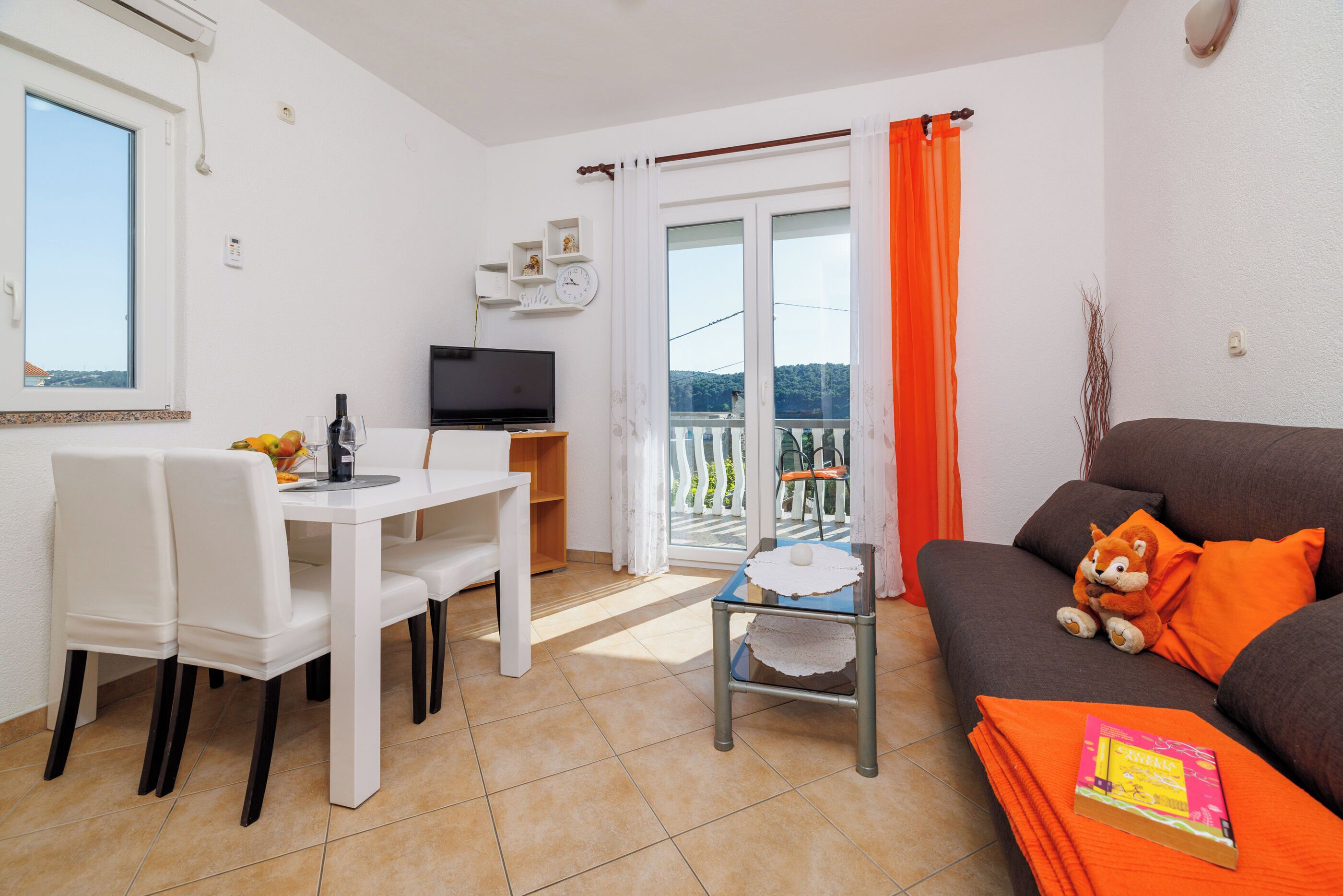 En stue med sofa, spisebord med stole, fjernsyn og en balkon med orange gardiner.