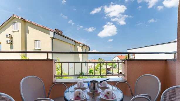 Un balcon avec une table ronde dressée pour le thé, entouré de chaises en métal, donnant sur un quartier résidentiel avec des maisons et un ciel dégagé.