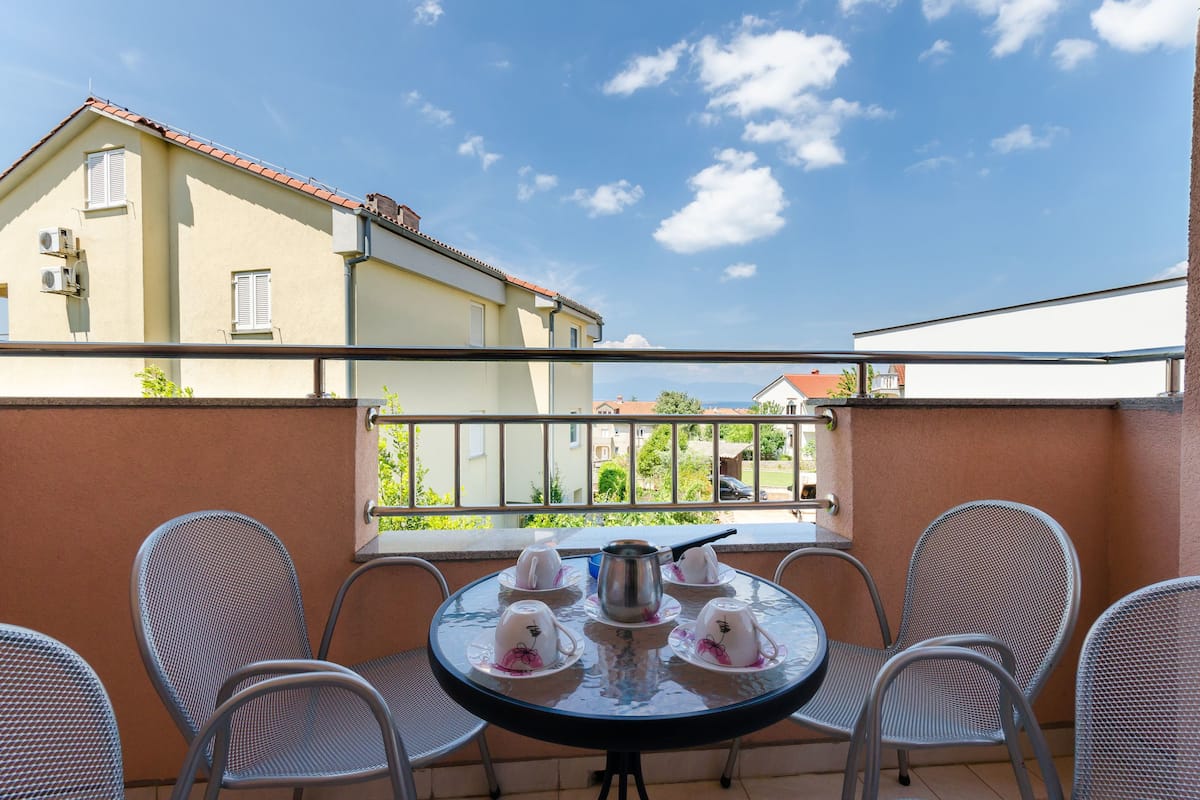 Ein Balkon mit rundem Tisch, gedeckt für Tee, umgeben von Metallstühlen, mit Blick auf ein Wohngebiet mit Häusern und einem wolkenlosen Himmel.