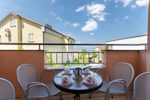 A balcony with a round table set for tea, surrounded by metal chairs, overlooking a residential area with houses and a clear sky. - Apartments Dunja (Malinska)