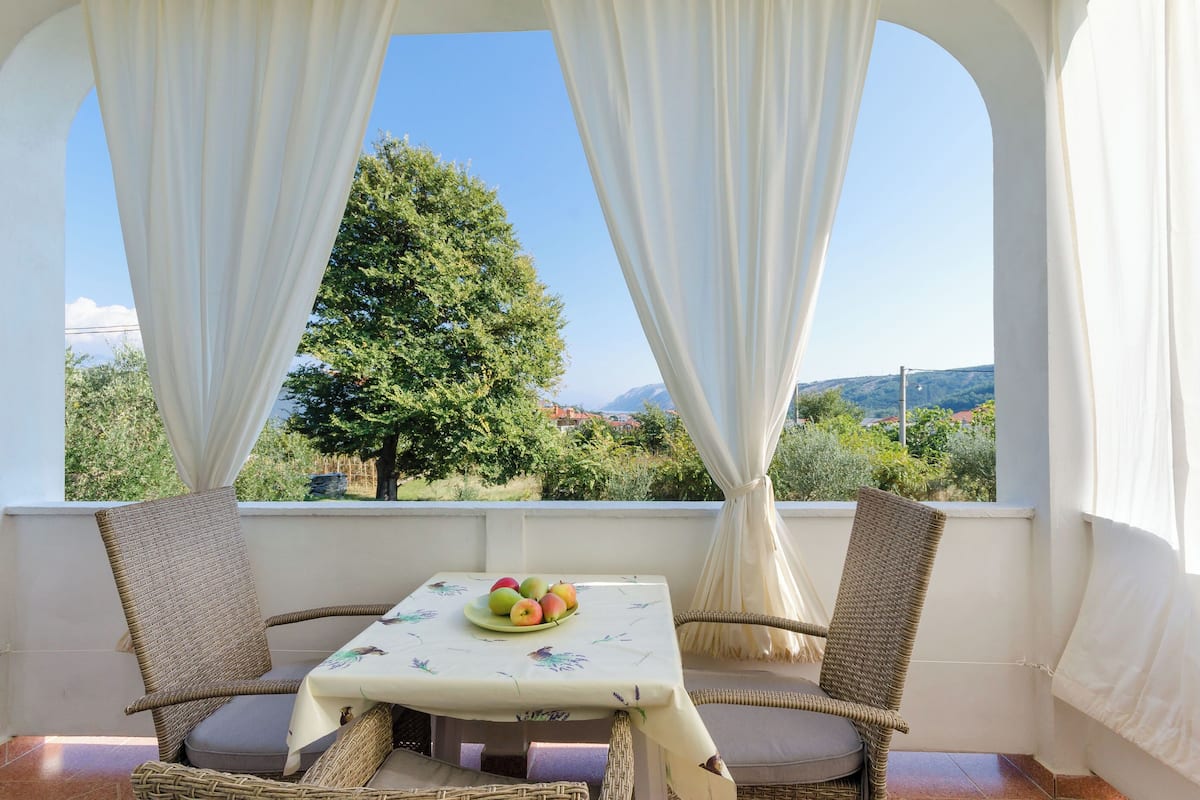 Un balcone con sedie in vimini, un tavolo con una ciotola di frutta e vista su un albero e sul paesaggio.