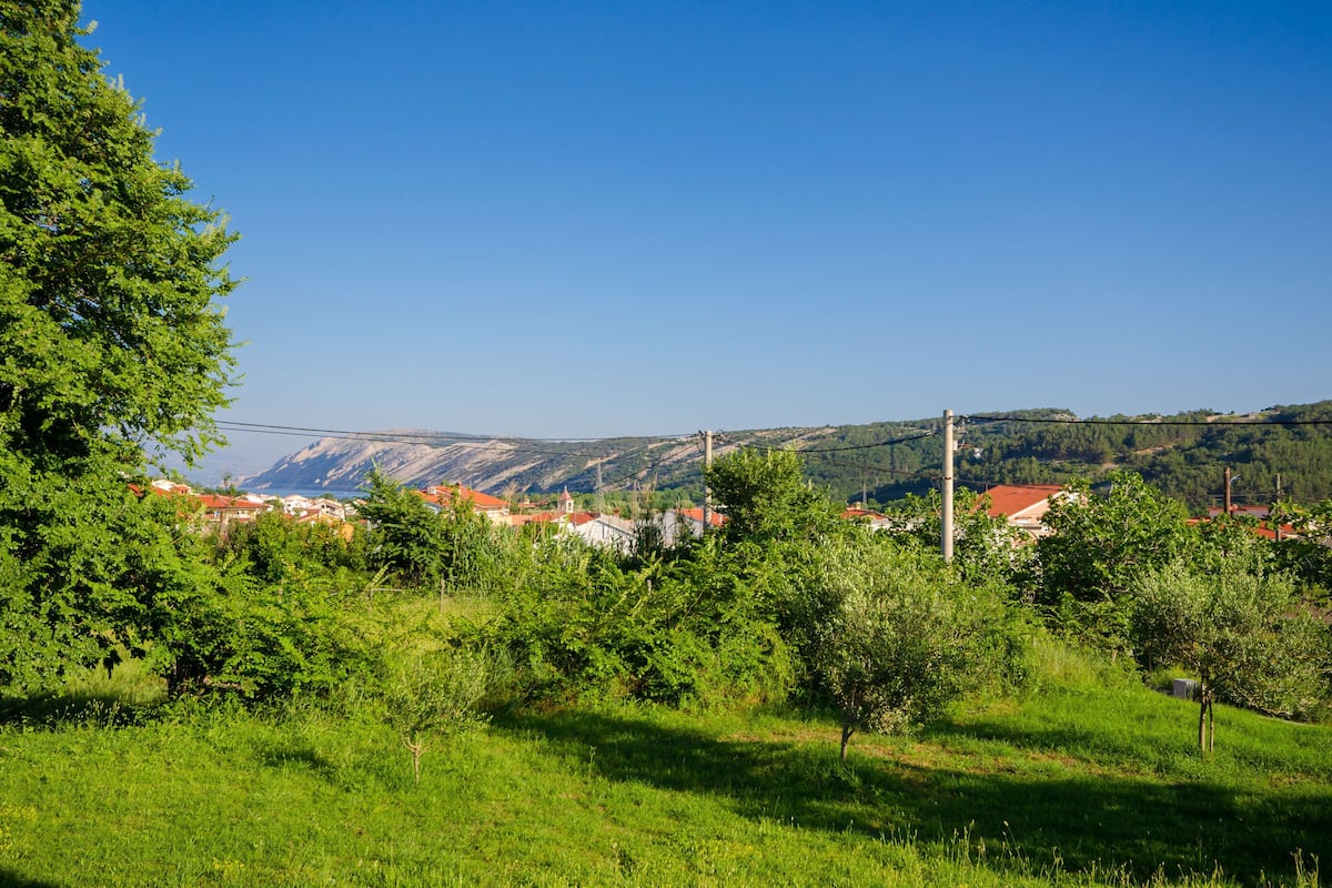 Paesaggio rurale con cielo sereno, campi verdi e un villaggio in lontananza con edifici dai tetti rossi.
