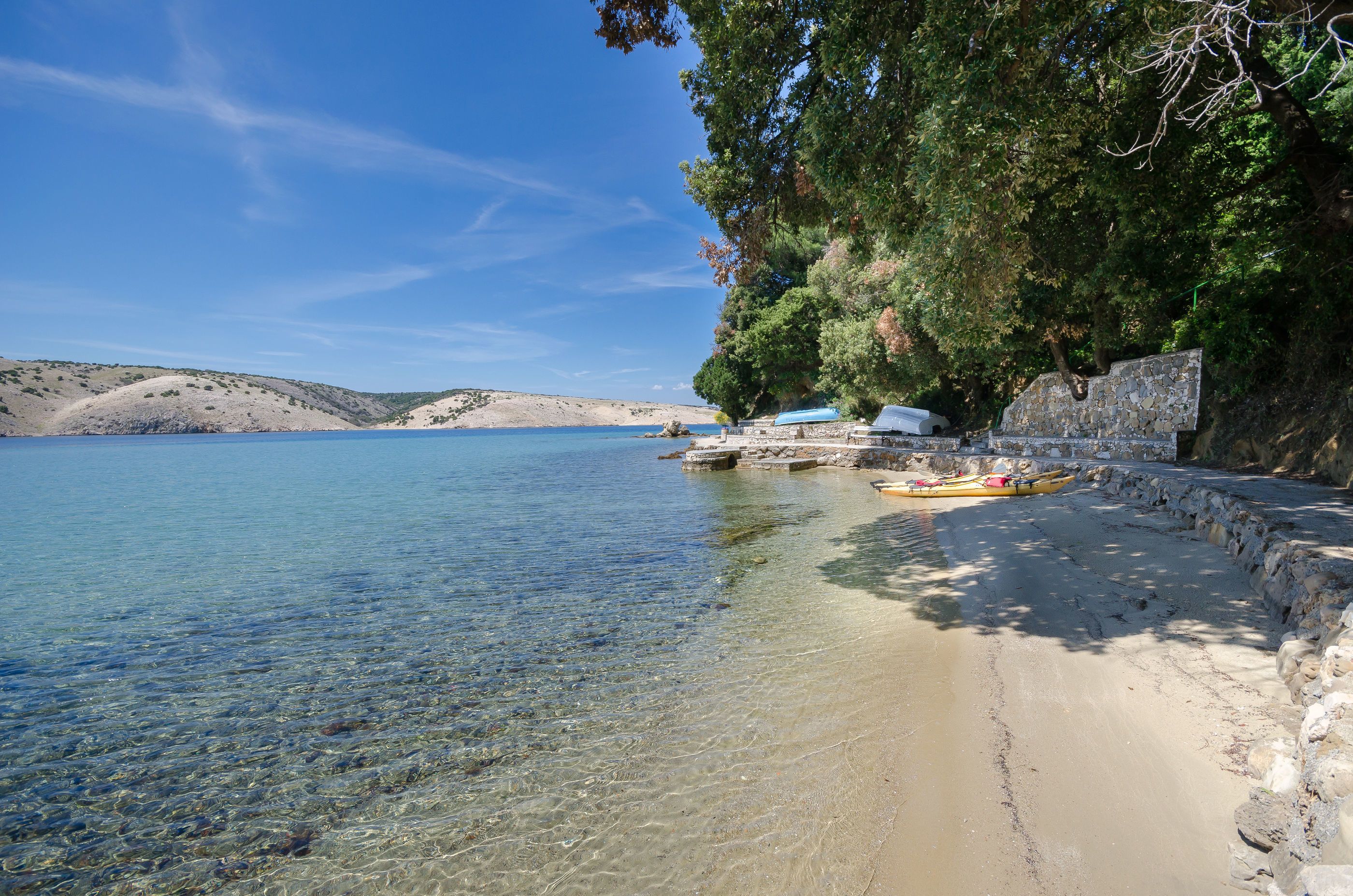 A beach with clear water, a stone wall, and a few boats.