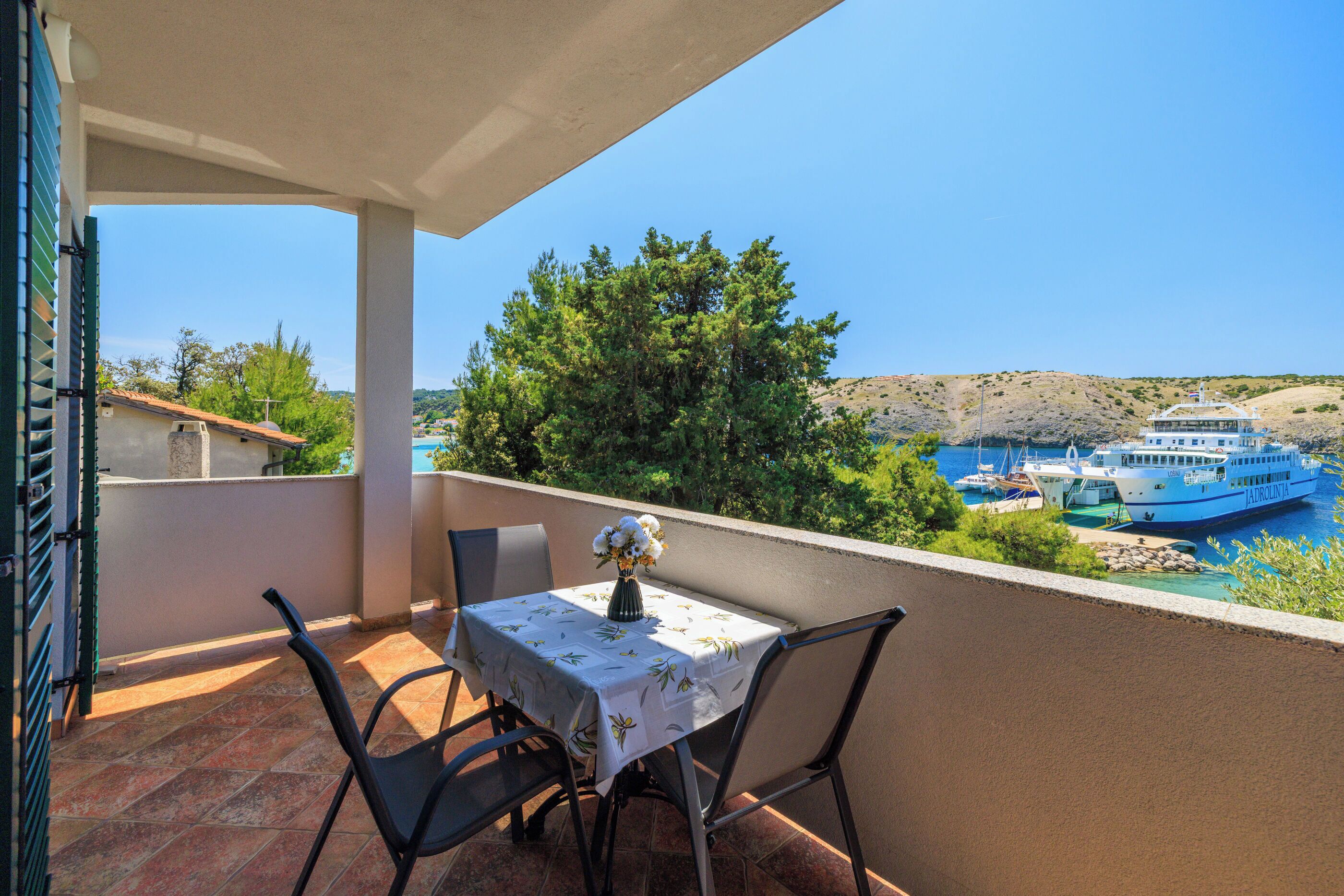 A balcony with a table and chairs, overlooking a body of water with boats and a hilly landscape.
