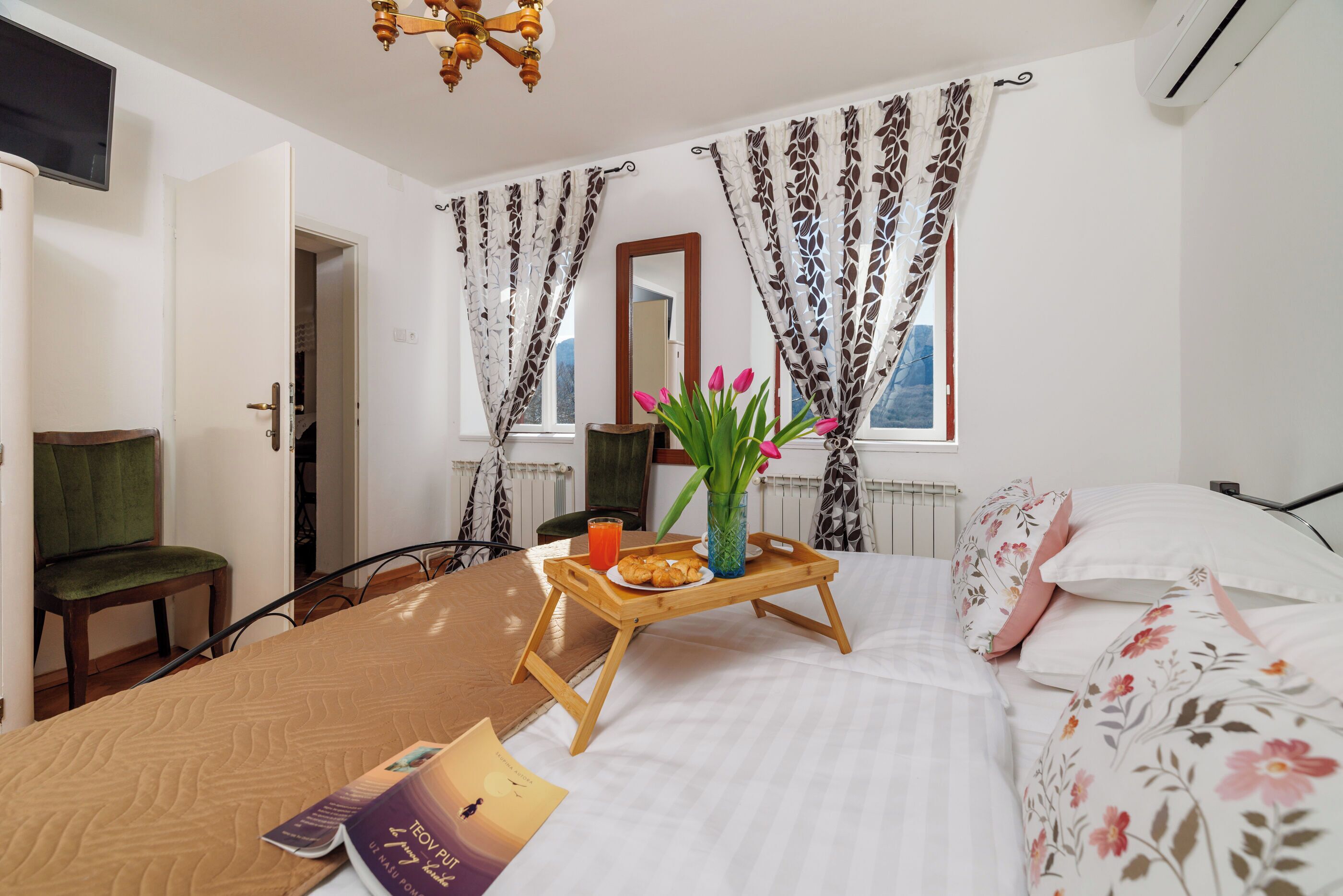 Ein Hotelzimmer mit Bett, einem Holftablett mit Frühstücksangeboten, einem grünen Sessel, einem Fenster mit Vorhängen und einem Spiegel.