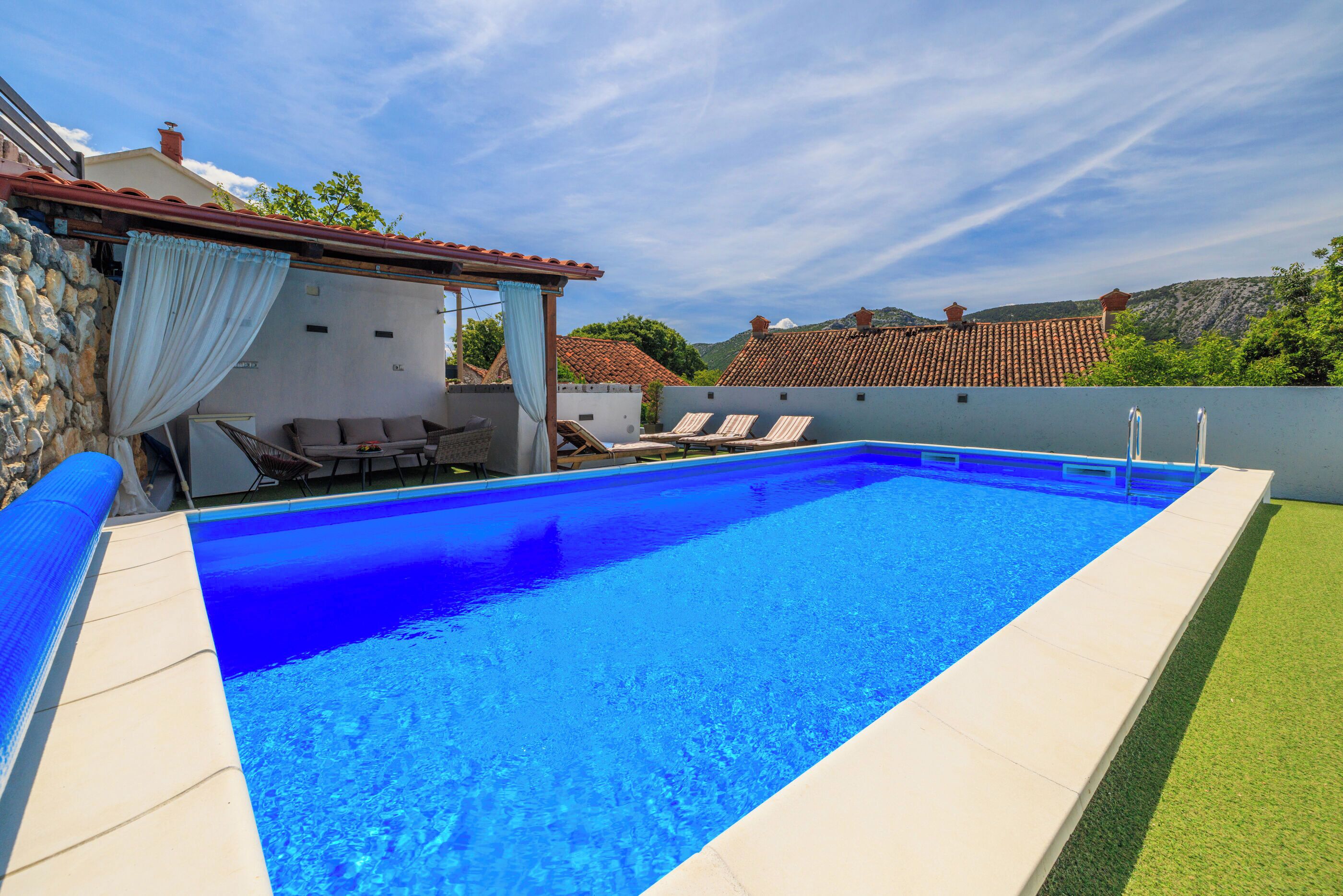A swimming pool with a clear blue water, surrounded by a white wall and a stone wall. There are lounge chairs and a table with a white tablecloth.