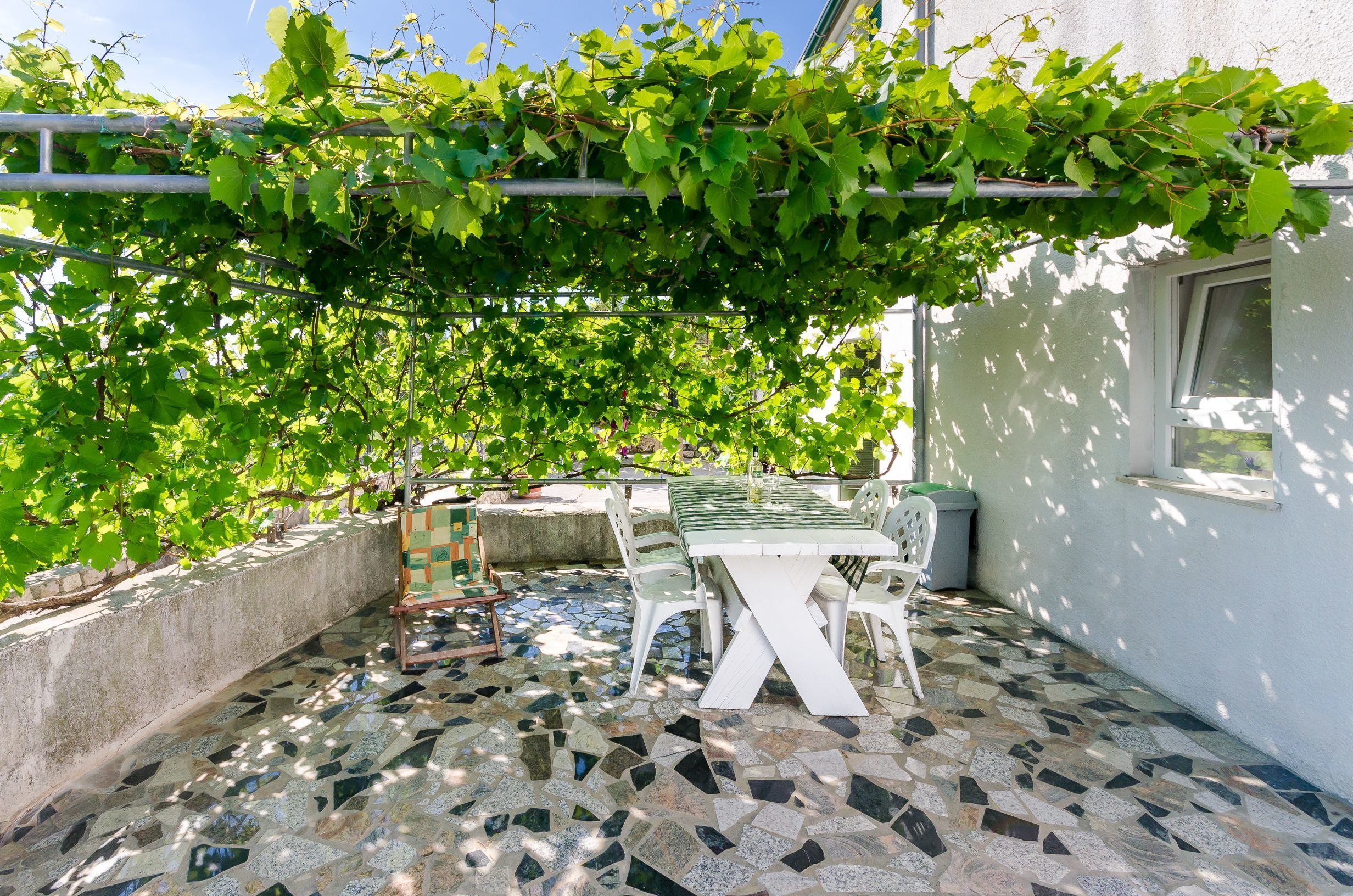 A patio with a mosaic floor, a white table and chairs, and a pergola covered with green vines.