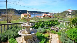 A garden with a stone fountain, a wooden fence, and a view of houses and hills in the background. - House Branka (Supetarska Draga)