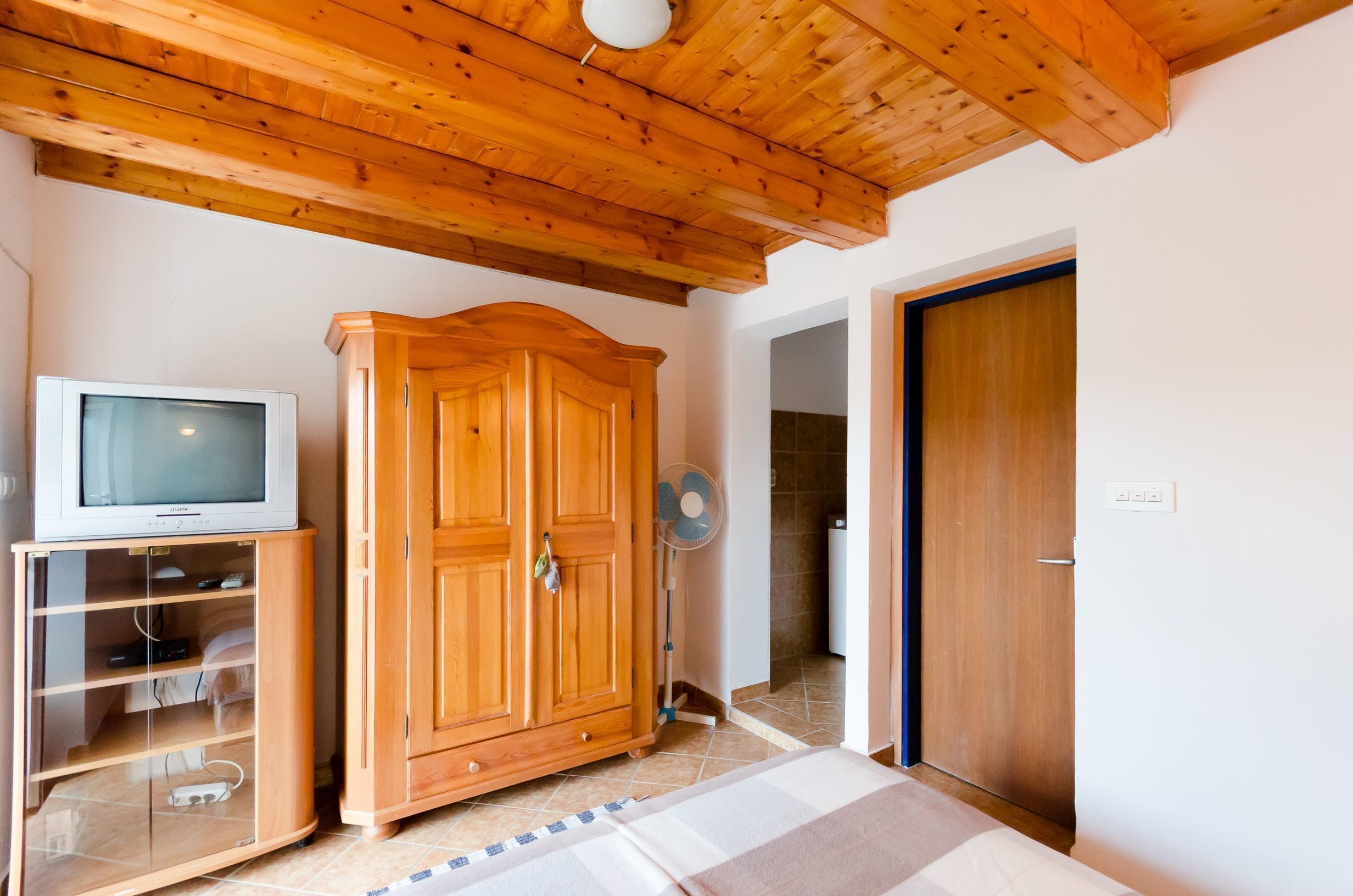 A room with a wooden ceiling, a wardrobe, a television on a stand, and a fan.