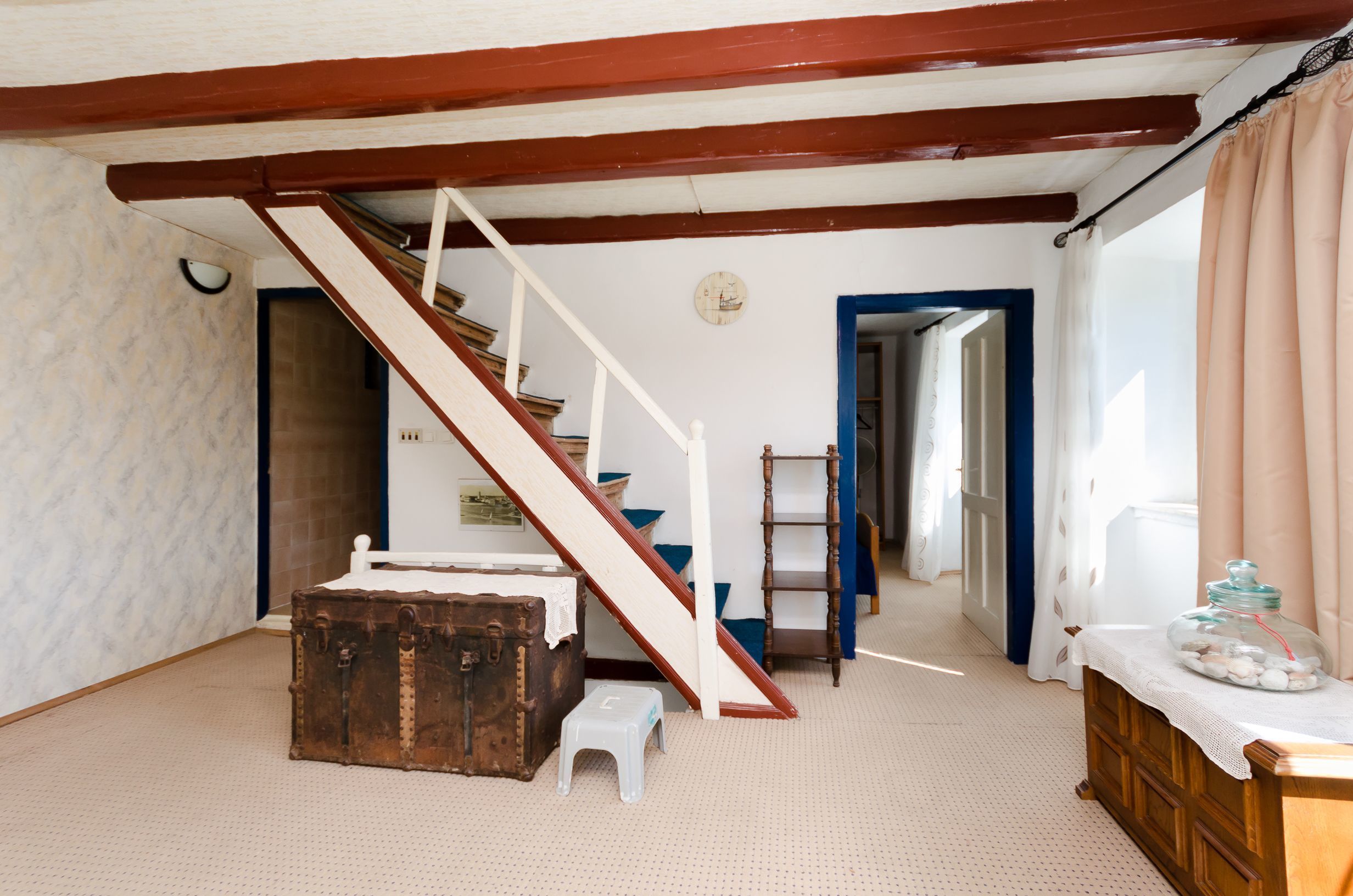 A room with a staircase, a wooden chest, a small stool, a ladder, and a window with curtains.