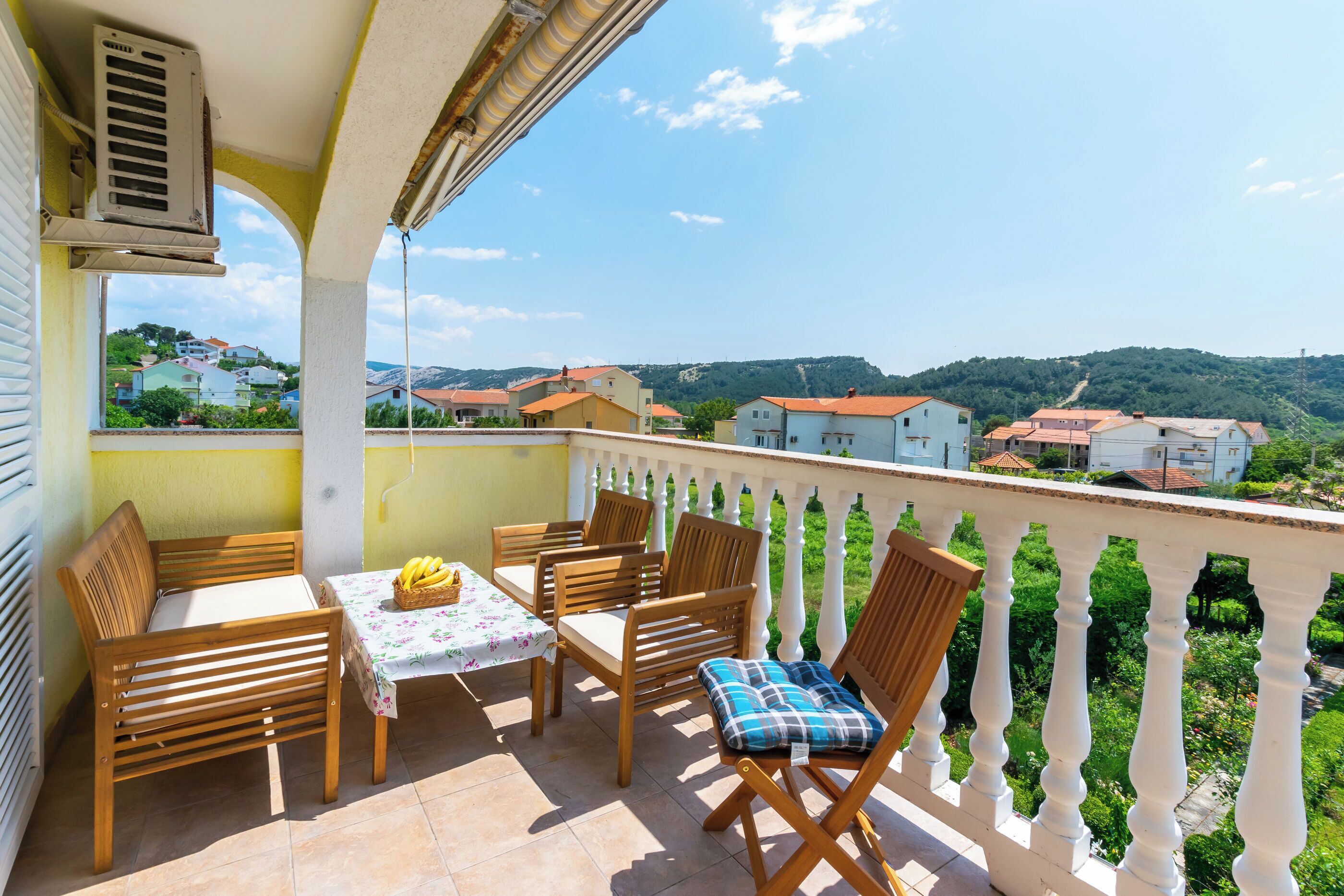 A balcony with wooden furniture, a table with a floral tablecloth, and a view of a hillside with buildings.