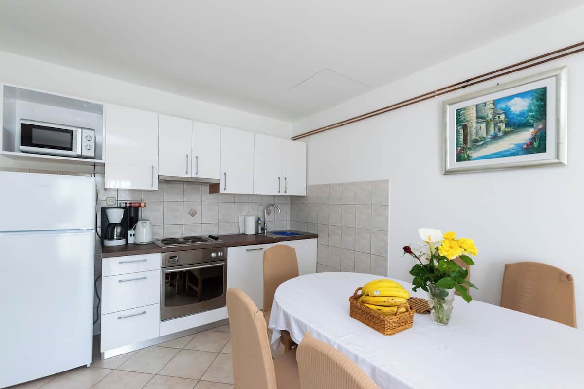 Una cocina moderna con armarios blancos, un microondas, una cafetera y un lavamanos. Hay una mesa de comedor con mantel blanco, sillas y un florero con flores amarillas.