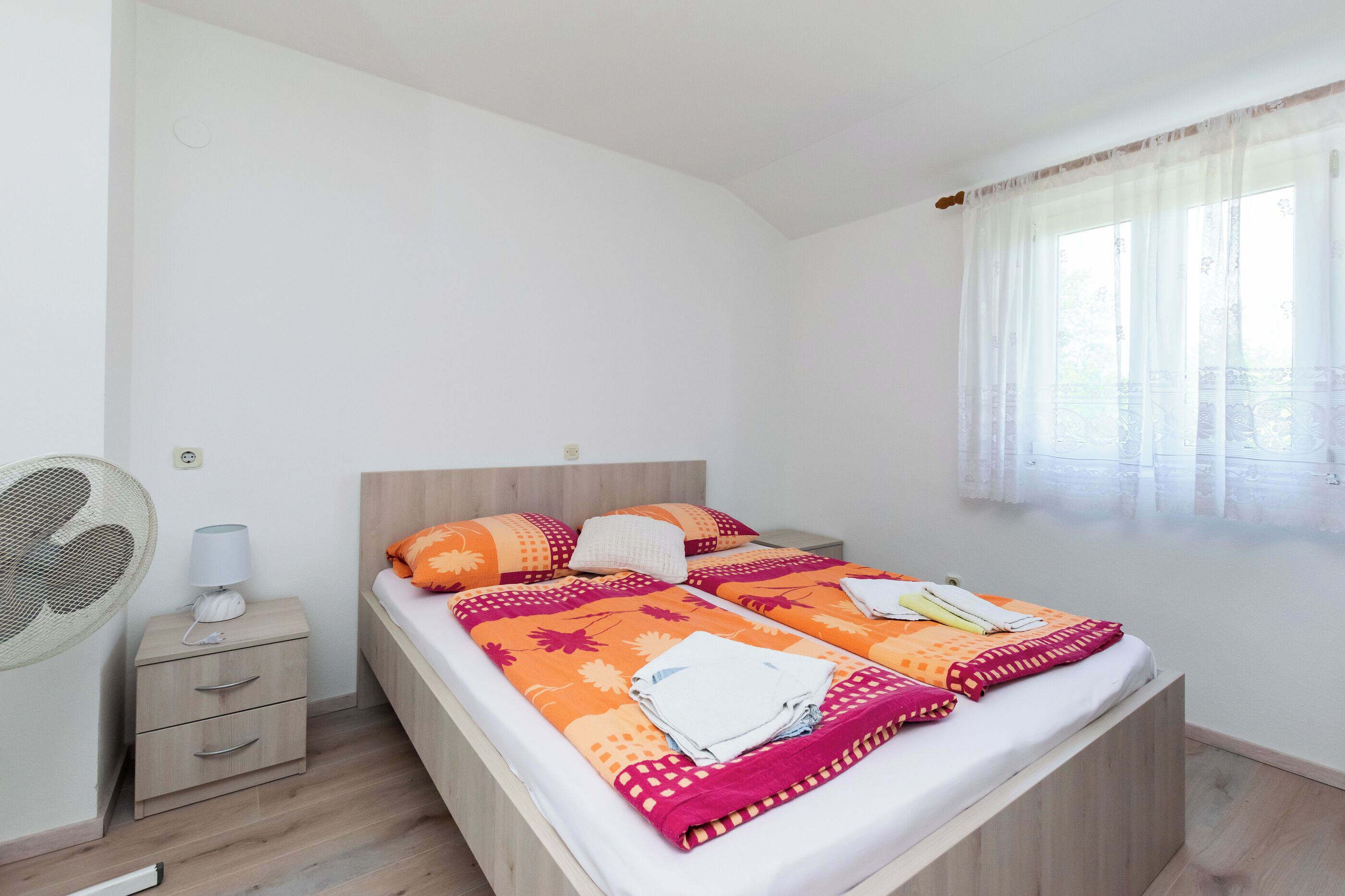 Ein Doppelbett mit orange-pink gestreifter Bettdecke, ein Nachttisch mit Lampe und ein Fenster mit transparenten Vorhängen.