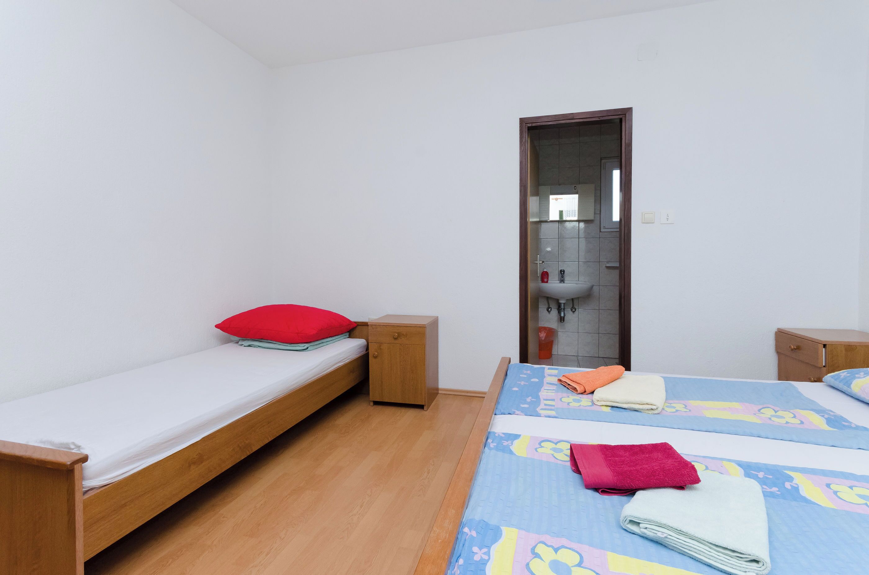 Ein kleines, sauberes Zimmer mit zwei Einzelbetten, einem hölzernen Nachttisch, einem roten Kopfkissen und einem gefalteten Handtuch auf einem Bett.