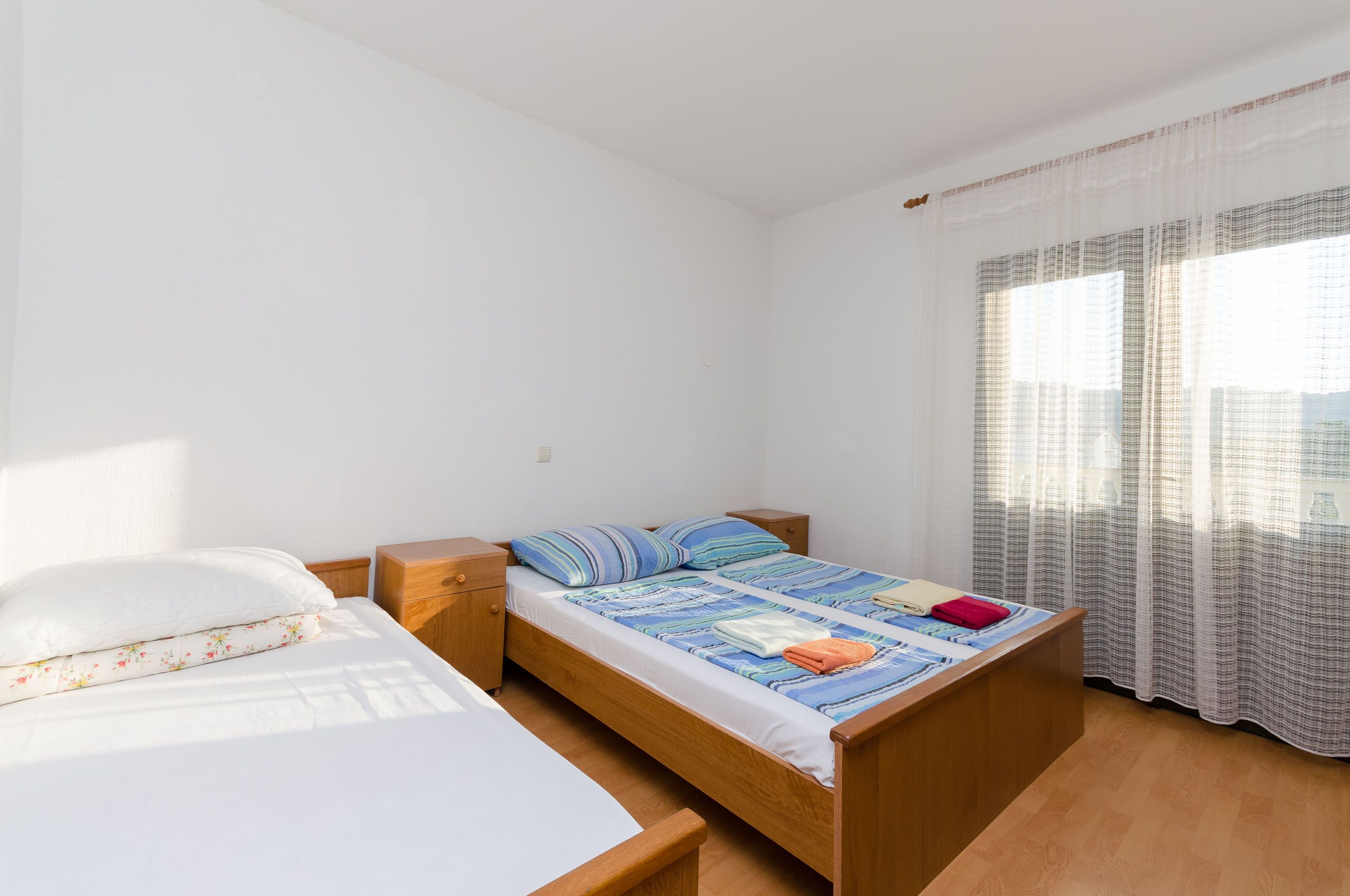 A single-bed room with a wooden frame, a white mattress, and a striped blanket. There is a wooden nightstand on one side and a window with sheer curtains.