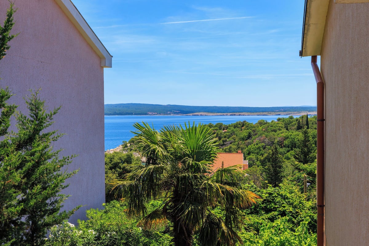 A view from a balcony overlooking a coastal landscape with a clear sky, a body of water, and lush greenery.
