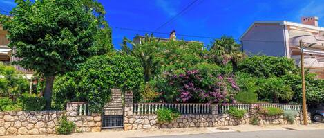 A residential street with a stone wall, a metal gate, and lush greenery including trees and flowering plants.