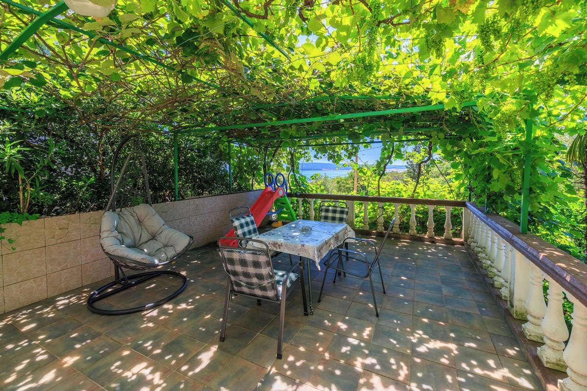 An outdoor patio with a table and chairs, surrounded by greenery and a view of the sea.