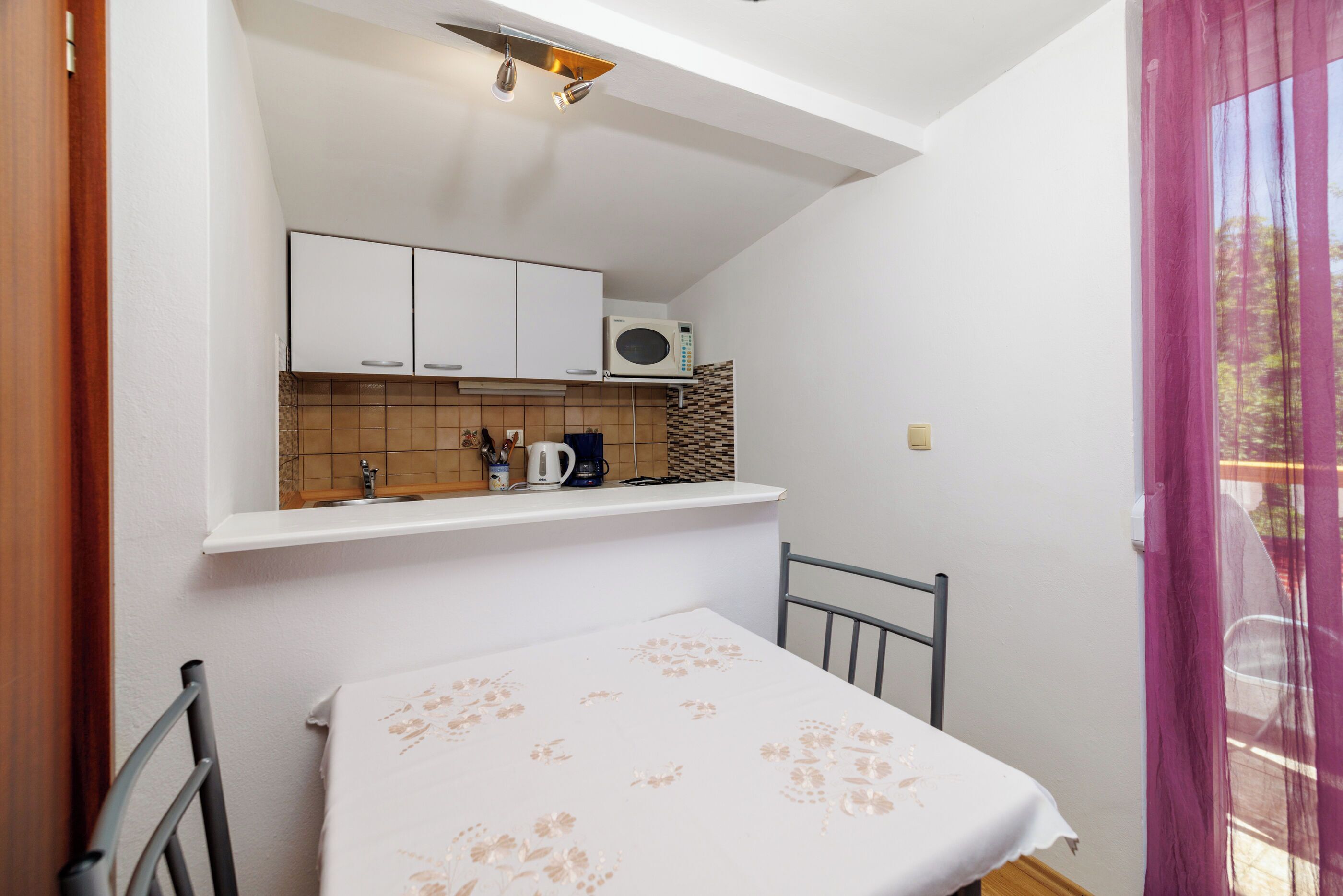 A small, compact room with a bed, a kitchenette with a microwave and cabinets, and a window with a sheer curtain.