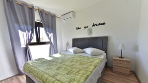 A bedroom with a bed, a bedside table with a lamp, a wall-mounted air conditioner, and a window with curtains. - Apartments Helena (Lopar)