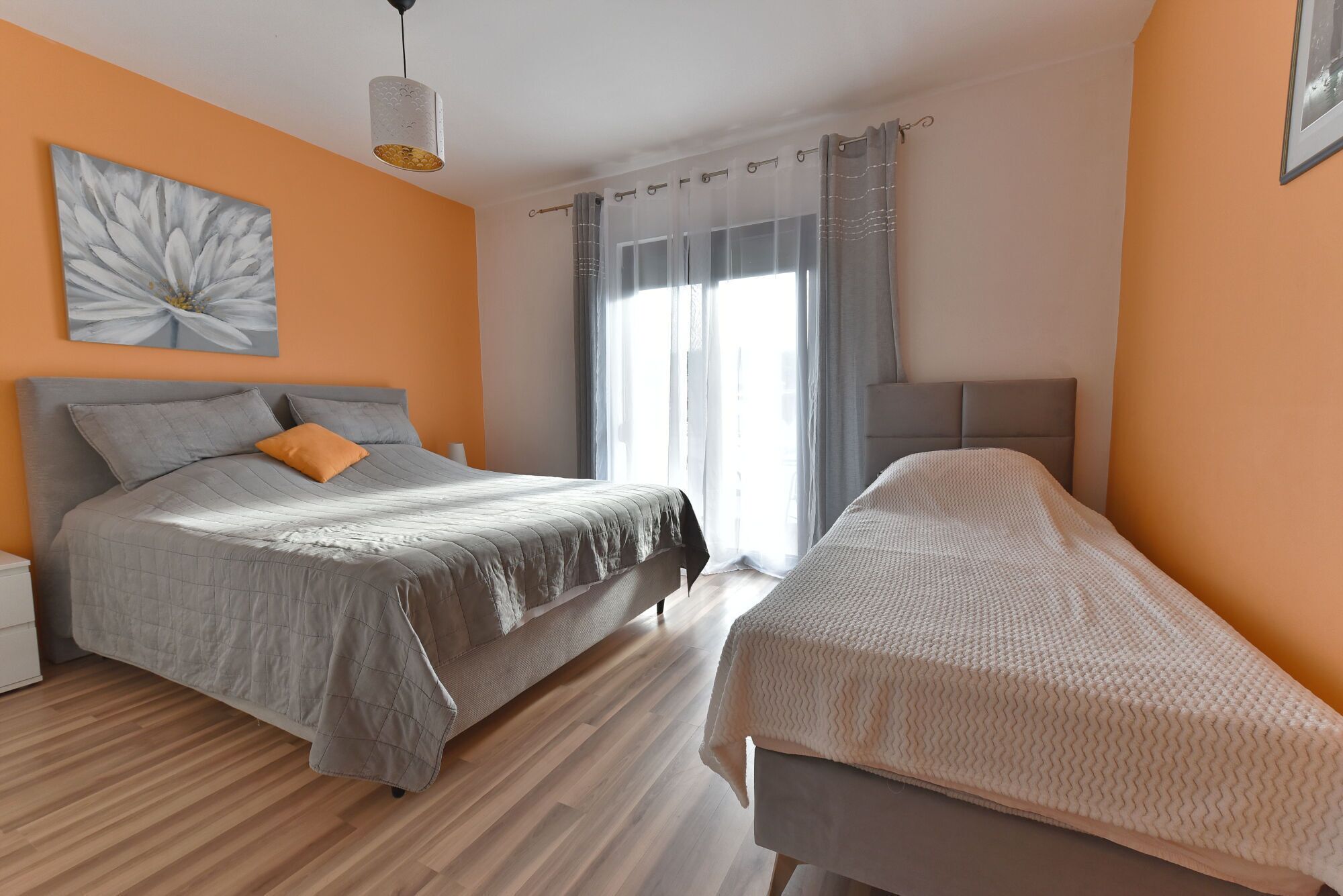 A bedroom with two beds, a large window with sheer curtains, and a wall-mounted light fixture.