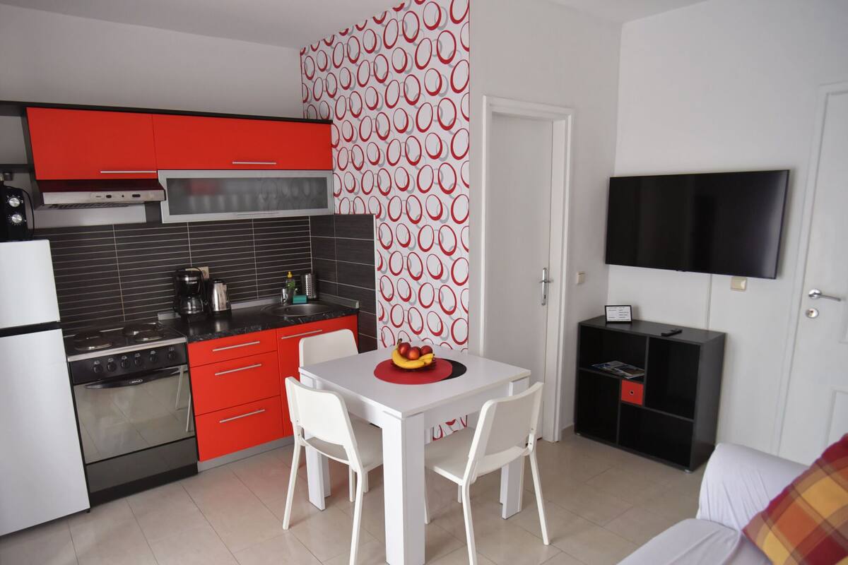 Una cucina compatta con elettrodomestici moderni, un'area pranzo con tavolo e sedie, e un televisore fissato alla parete.