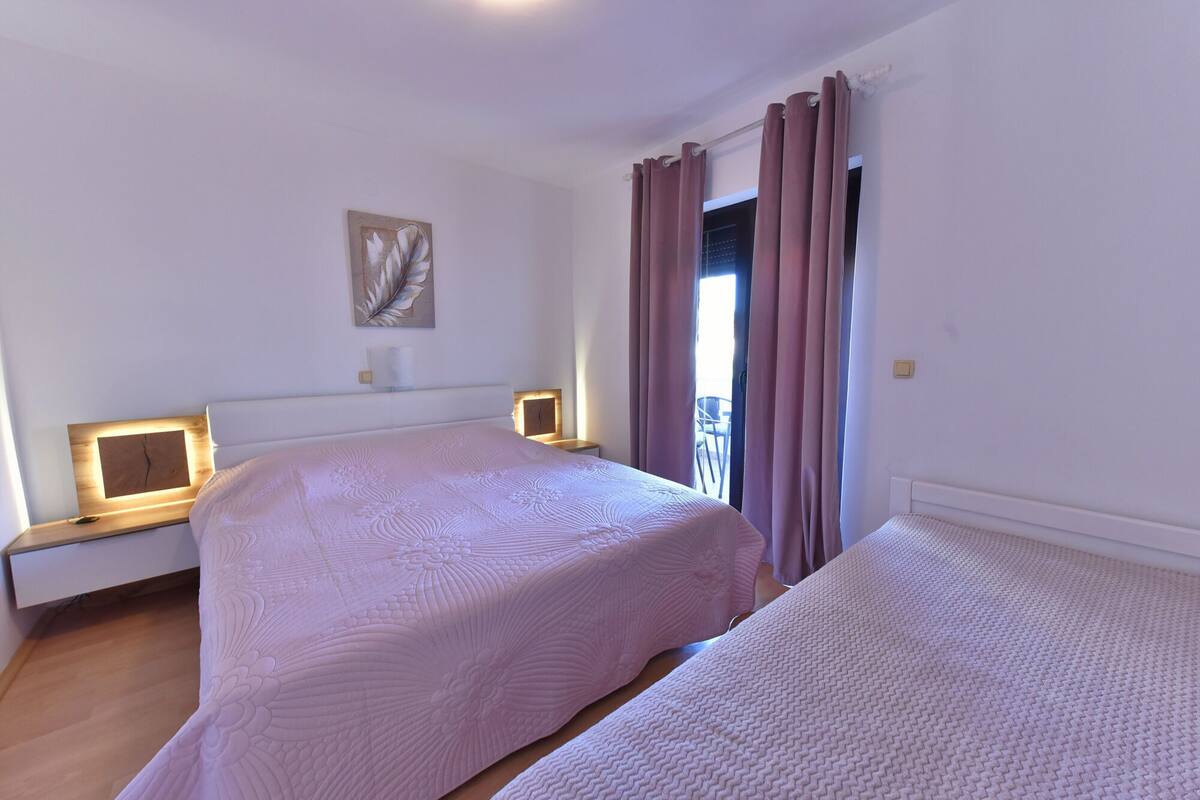Una camera d'albergo con due letti, un comodino con una lampada, un'opera d'arte appesa al muro e vista attraverso una finestra con tende.