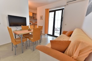 A living room with a sofa, a dining table with chairs, a flat-screen TV, and a kitchen area in the background. - Apartments Helena (Lopar)