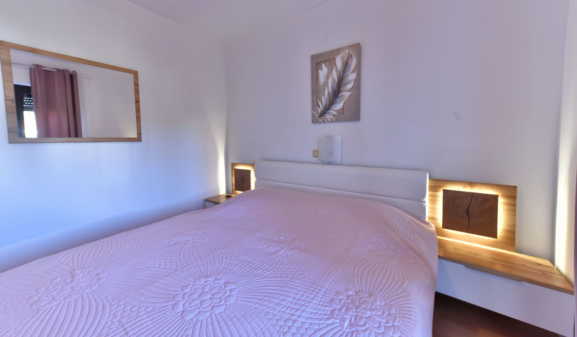 A bedroom with a large bed, a bedside table, a nightstand, a wall-mounted lamp, and a framed artwork.
