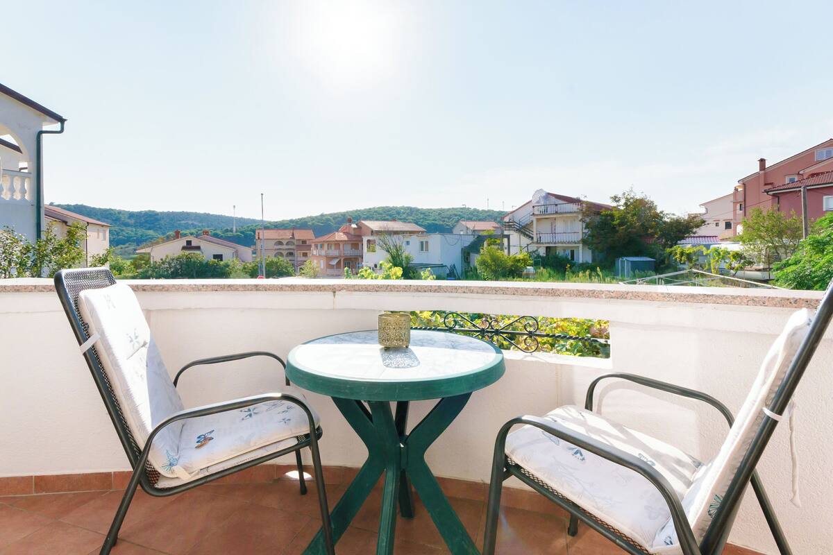 Un balcone con due sedie e un tavolino, affacciato su una zona residenziale con case e colline.
