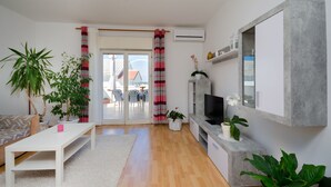 A modern living room with a white sofa, a coffee table, a TV, and a fireplace. There are potted plants and a large window with curtains. - Apartment Milja (Banjol)