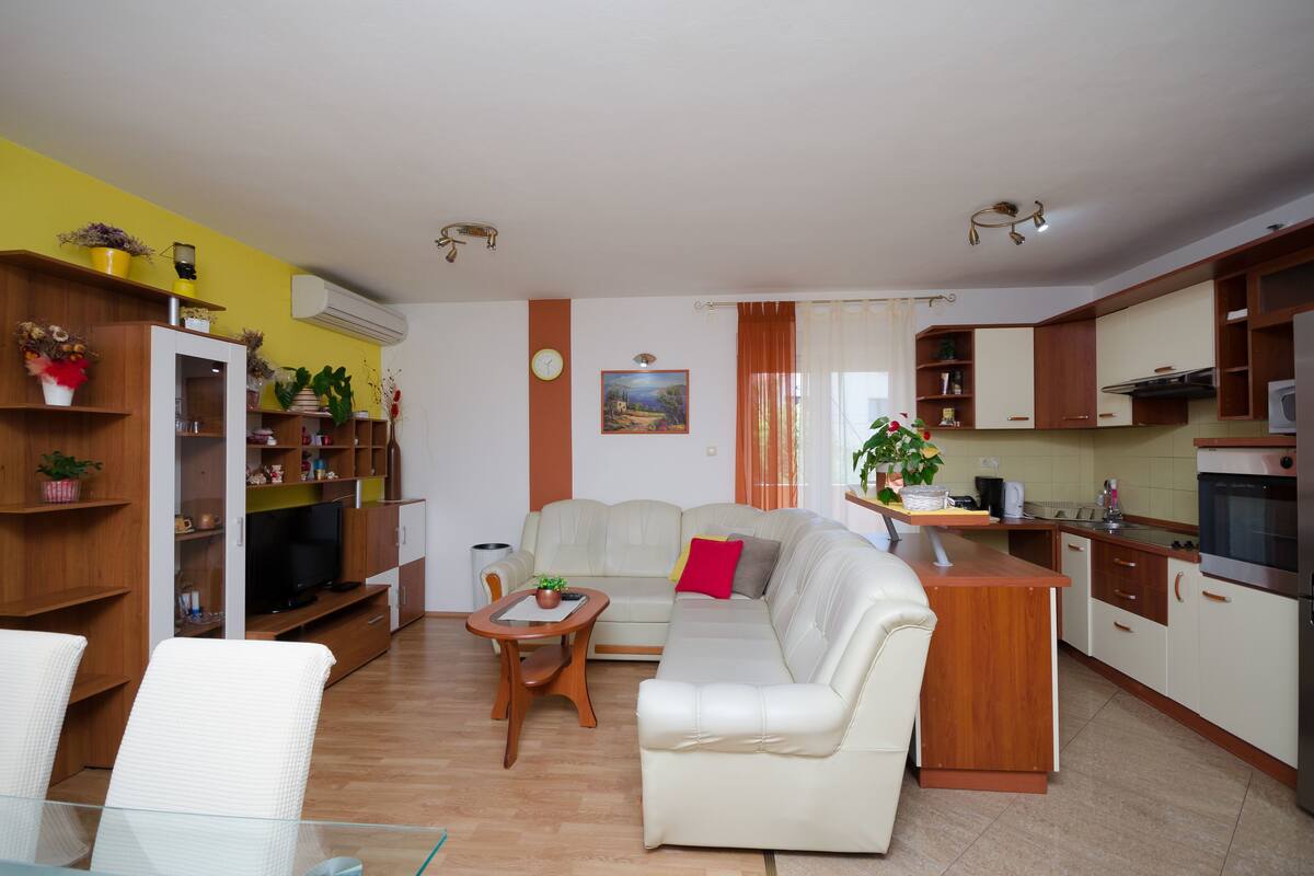 Una sala de estar moderna con sofá, mesa de centro y televisor. Se observa una cocina con armarios y un refrigerador.