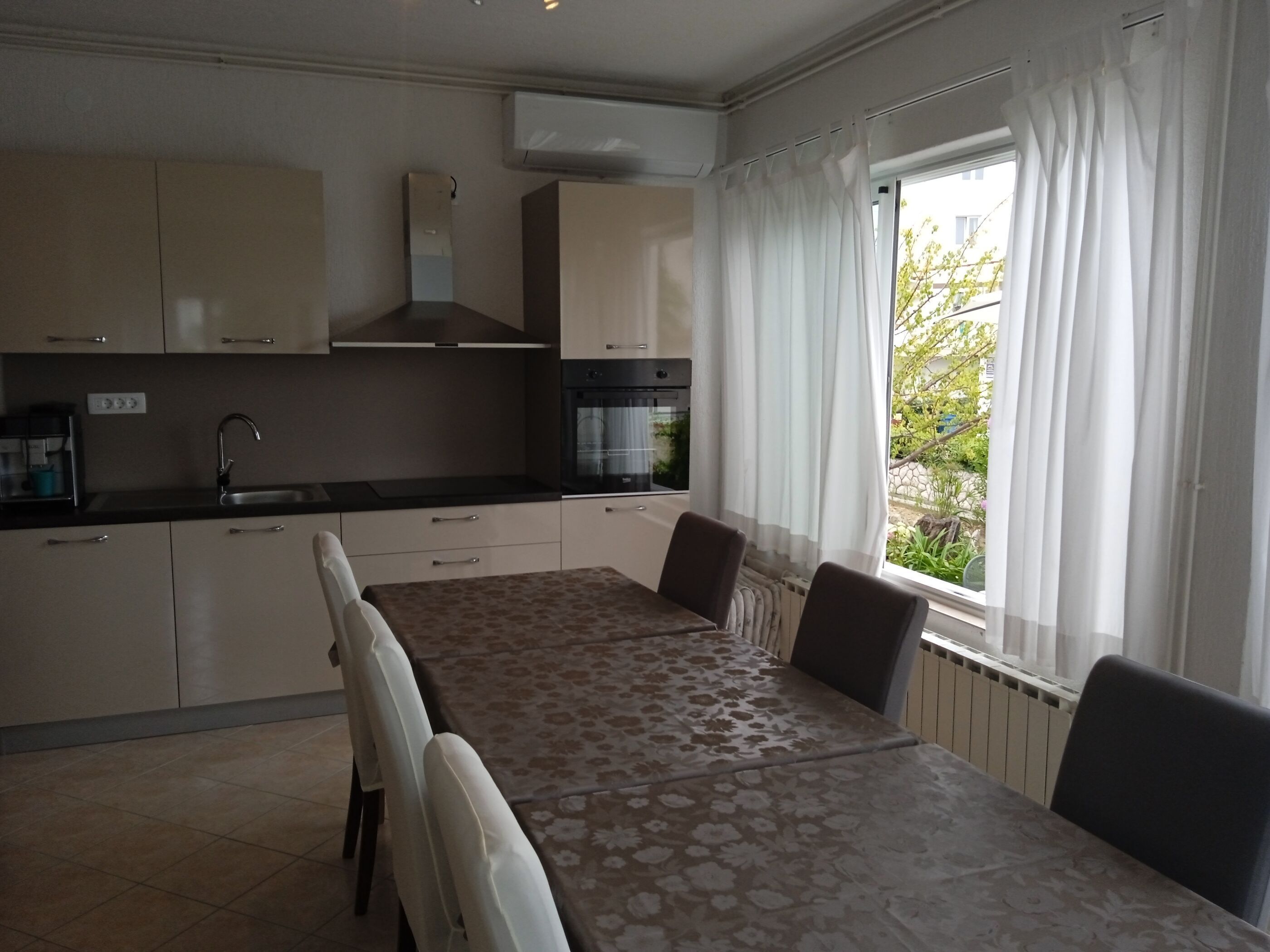 A modern kitchen with a dining table and chairs, a built-in oven, and a window with curtains.