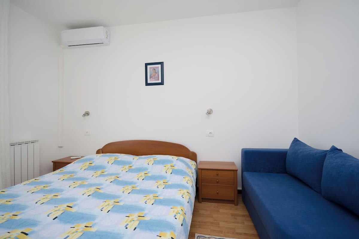 Ein Schlafzimmer mit einem Bett, einem blauen Sofa, einem Nachttisch, einem gerahmten Bild an der Wand und einer Klimaanlage.