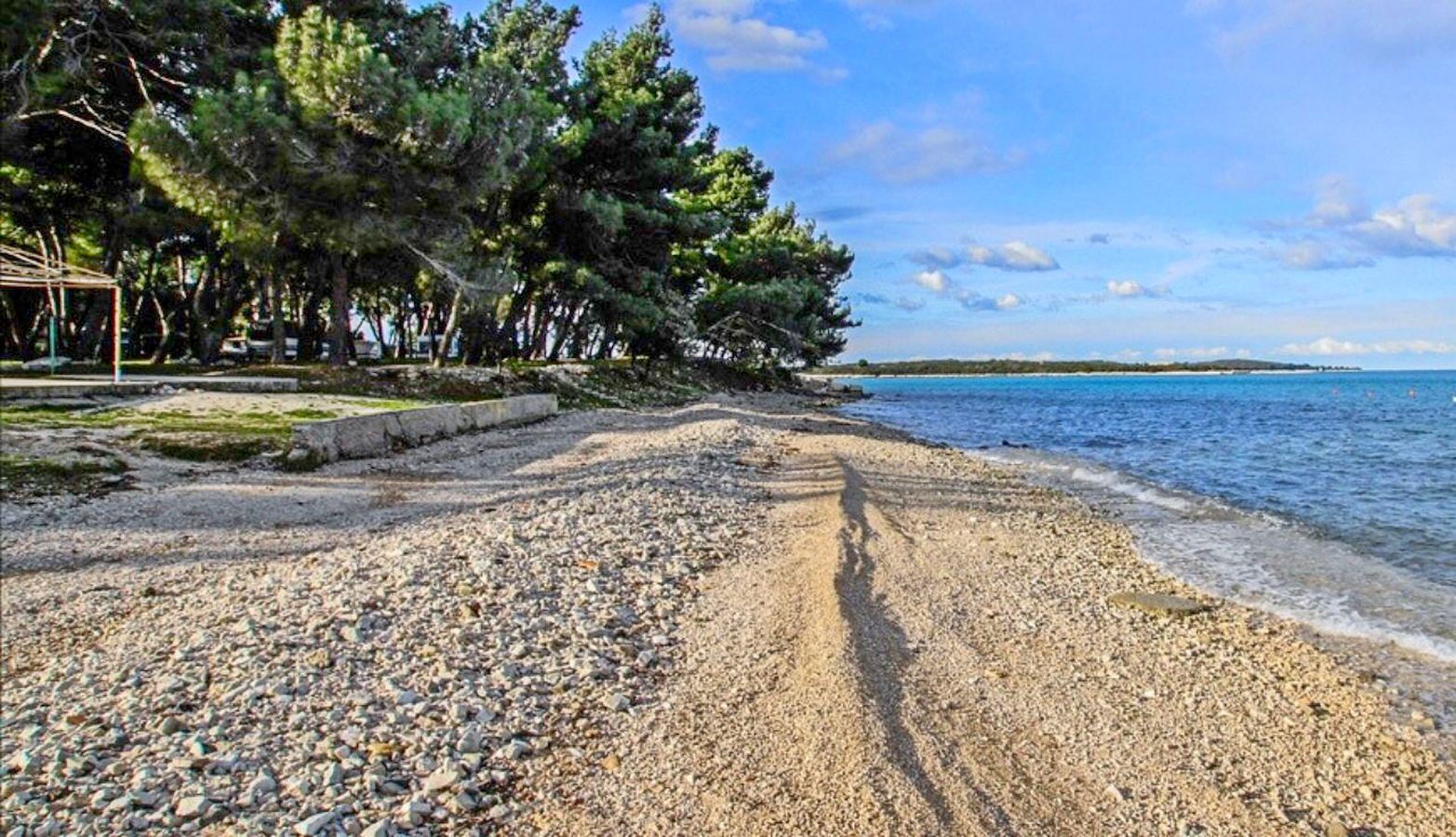 A pebbled beach with a clear view of the sea and a row of trees in the background.