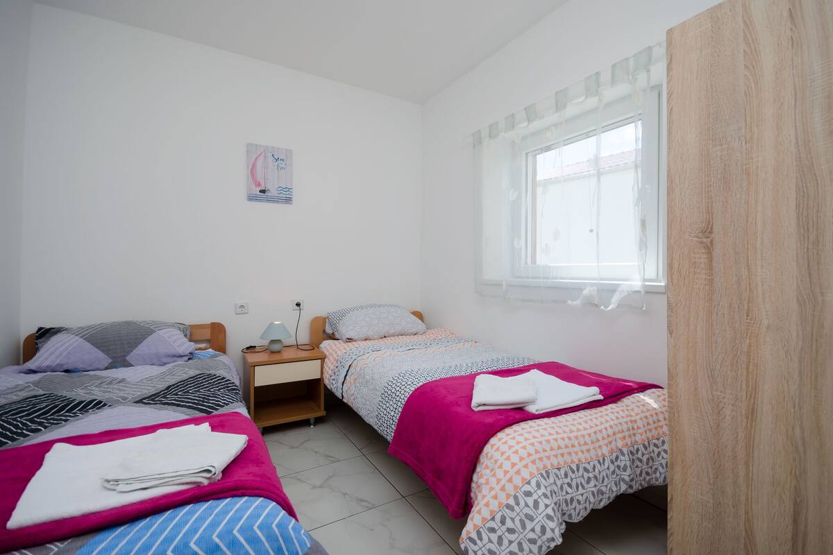 Ein kleines, sauberes Zimmer mit zwei Betten, einem Nachttisch und einem Fenster mit durchsichtigen Vorhängen.