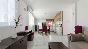 A modern living room with a sofa, a coffee table, and a dining area with chairs and a table. A kitchen is visible in the background with cabinets and appliances. - Apartments Slobodanka (Lopar)