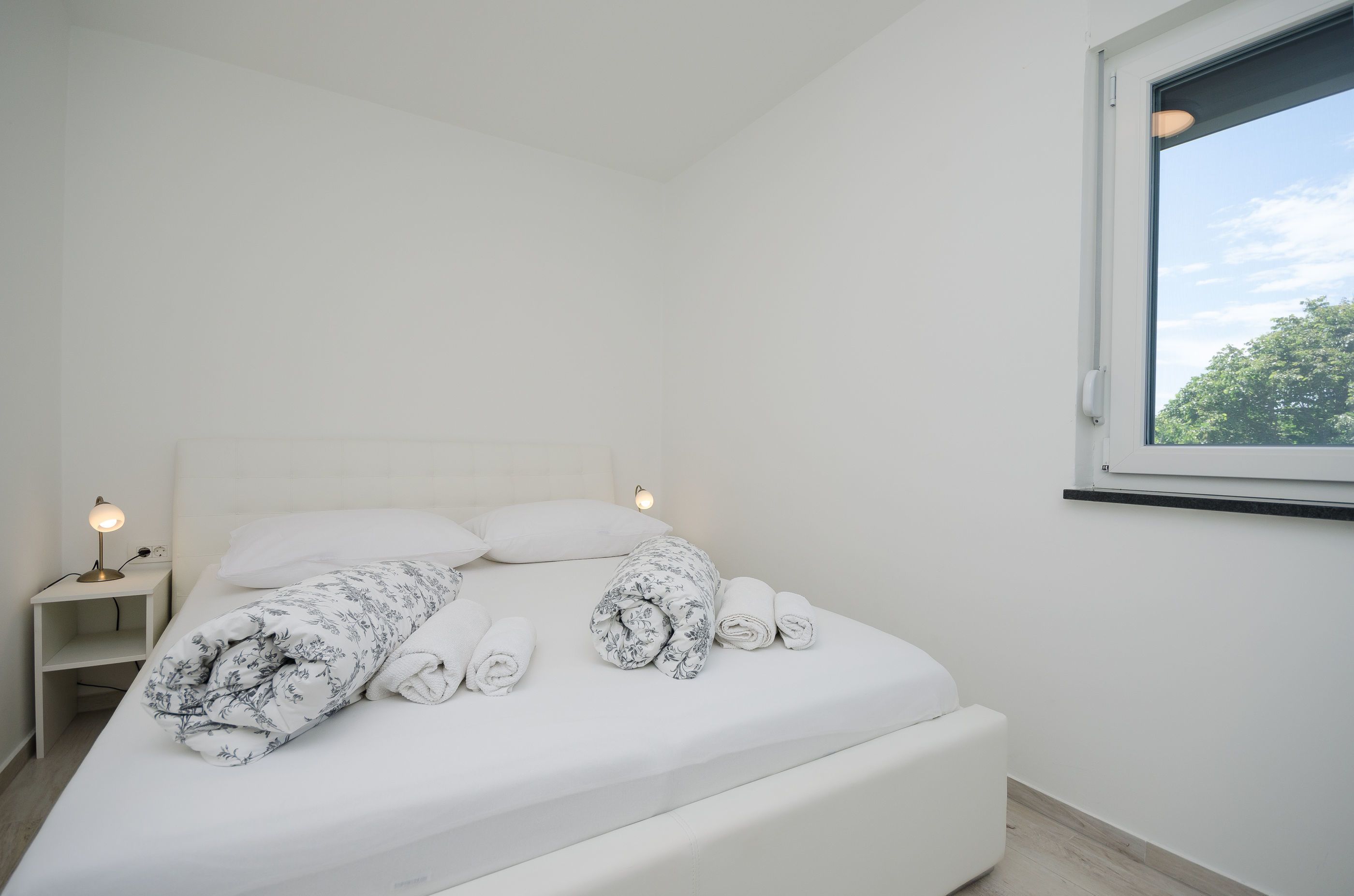 A neatly made bed with rolled towels, a bedside lamp, and a window with a view of trees.