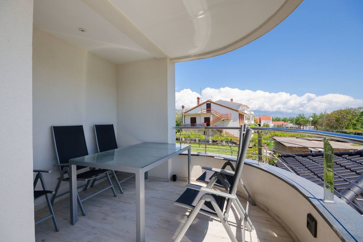 A balcony with a glass table and chairs, overlooking a residential area with houses and a clear sky.