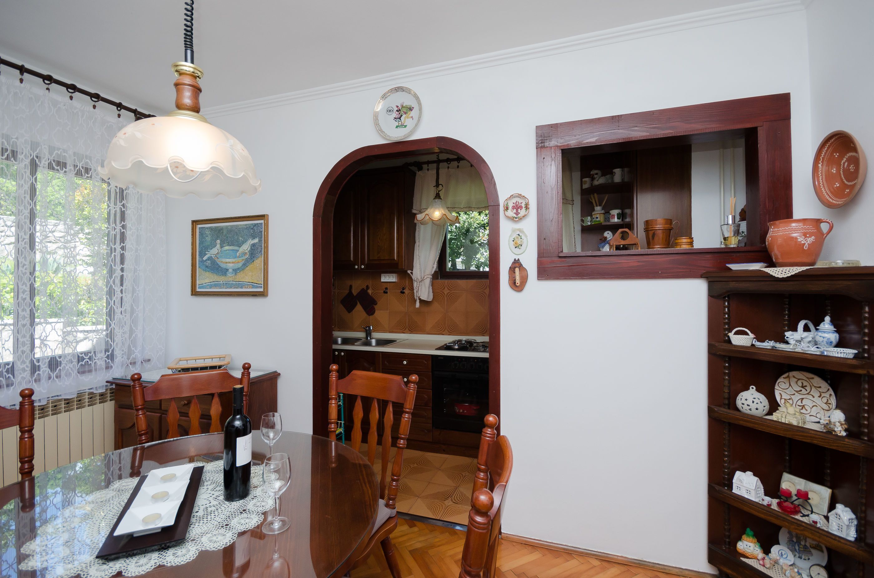 A dining area with a wooden table, chairs, and a chandelier. There is a kitchen visible through an archway.