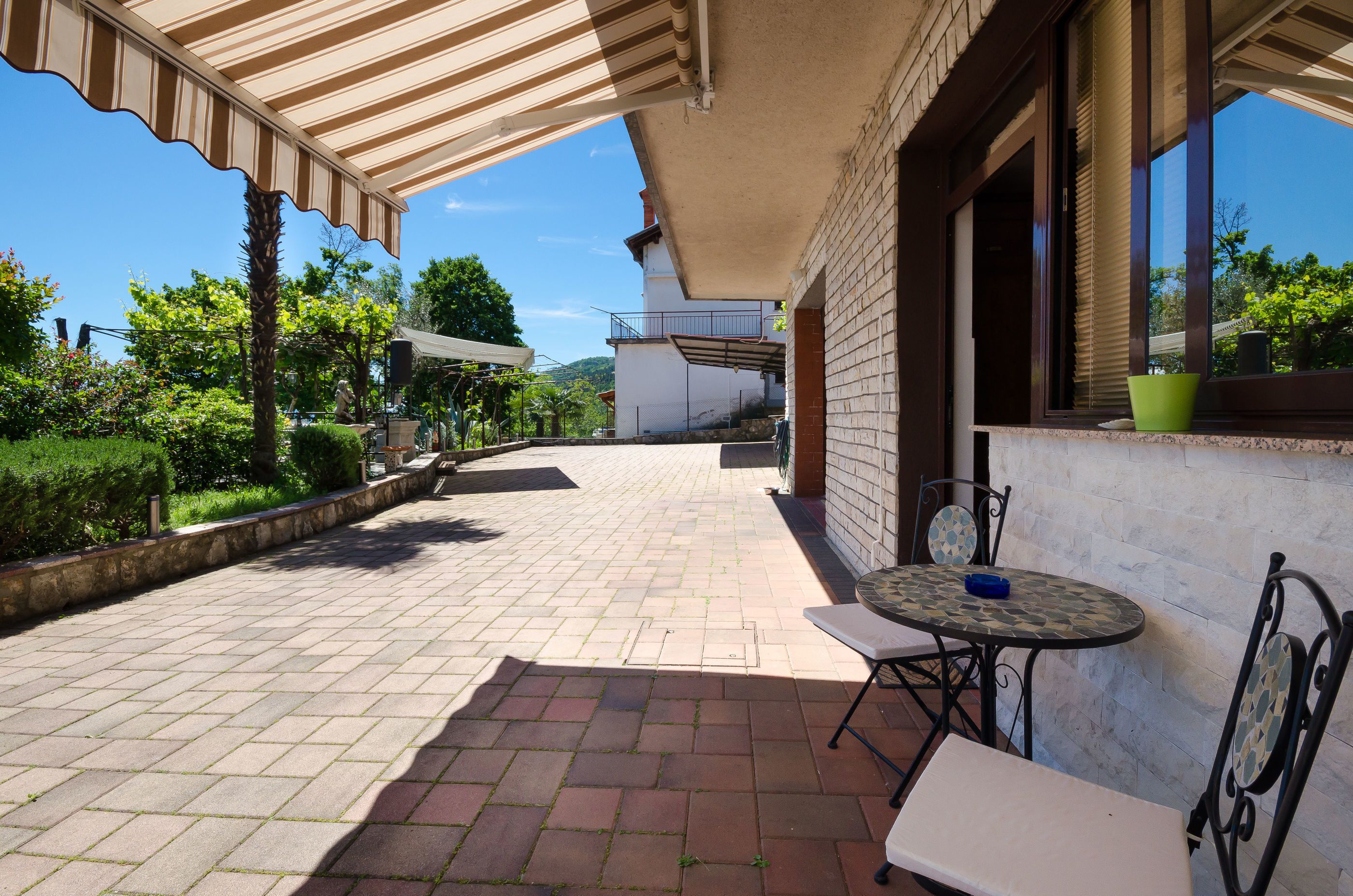 A covered patio area with a table and chairs, a brick pathway, and a view of greenery and buildings in the background.