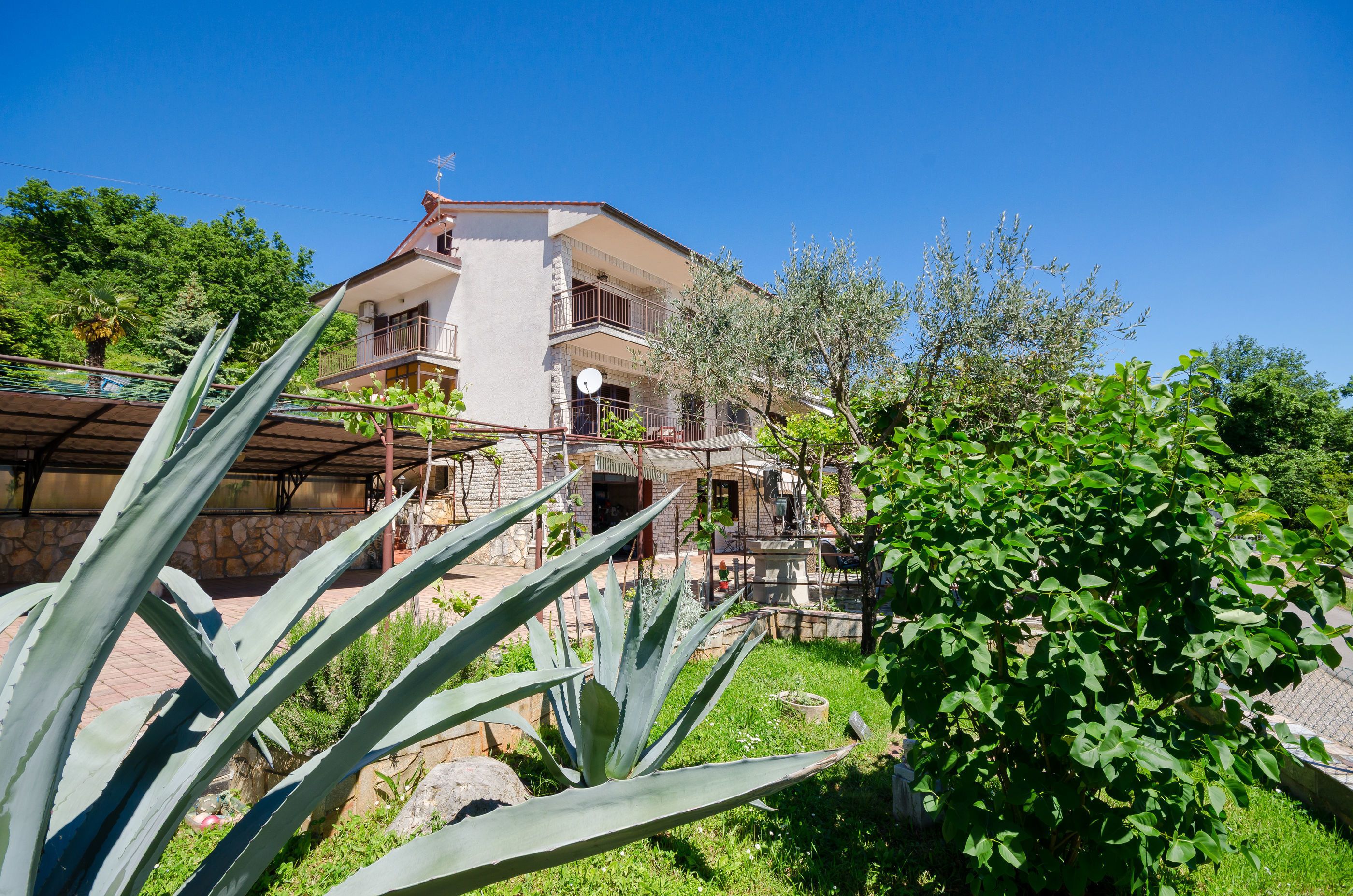 A two-story house with a balcony, surrounded by greenery and a clear blue sky.