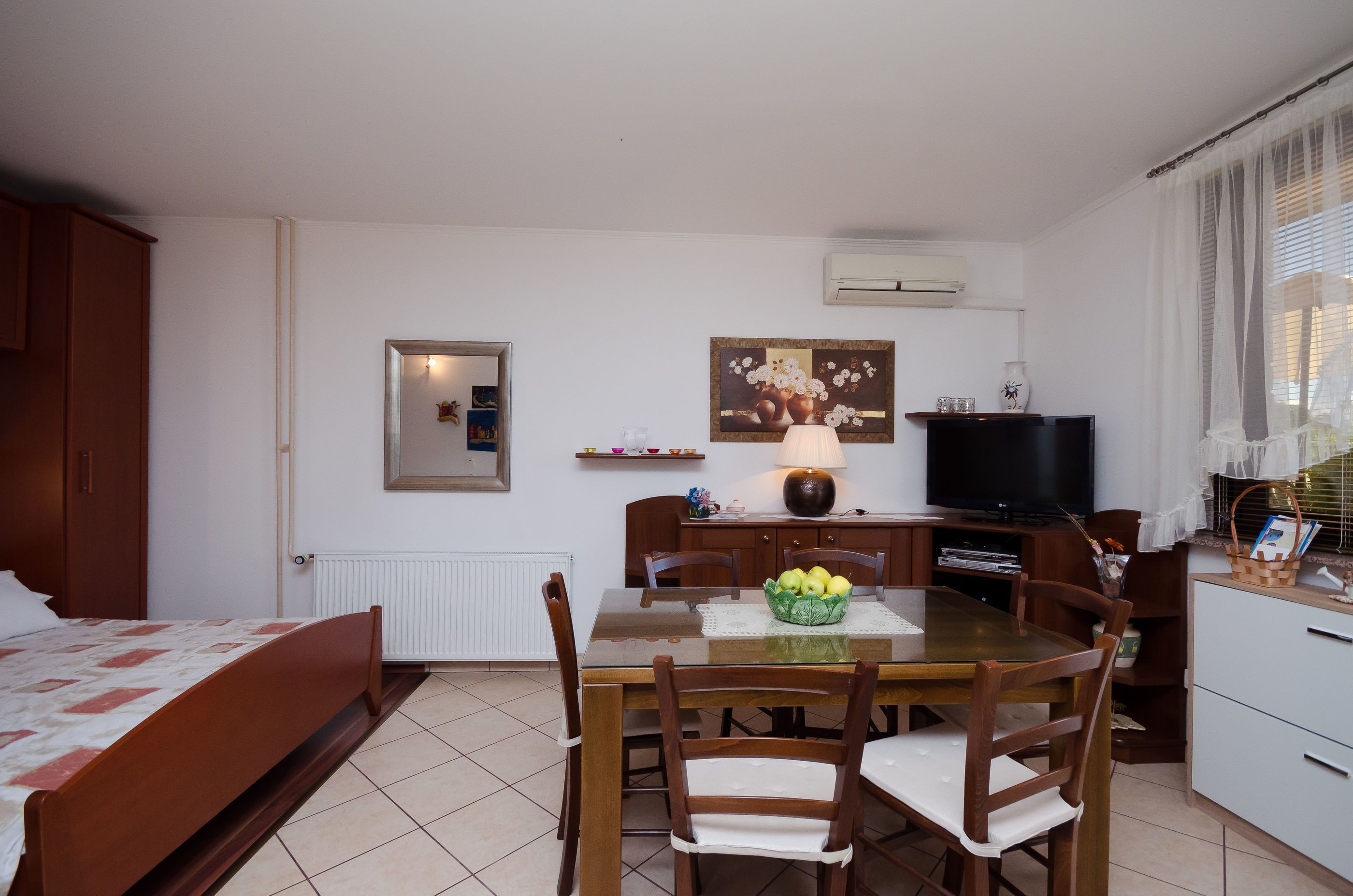 Un dormitorio con cama, una pequeña zona de comedor con mesa y sillas, un televisor y una ventana con cortinas.