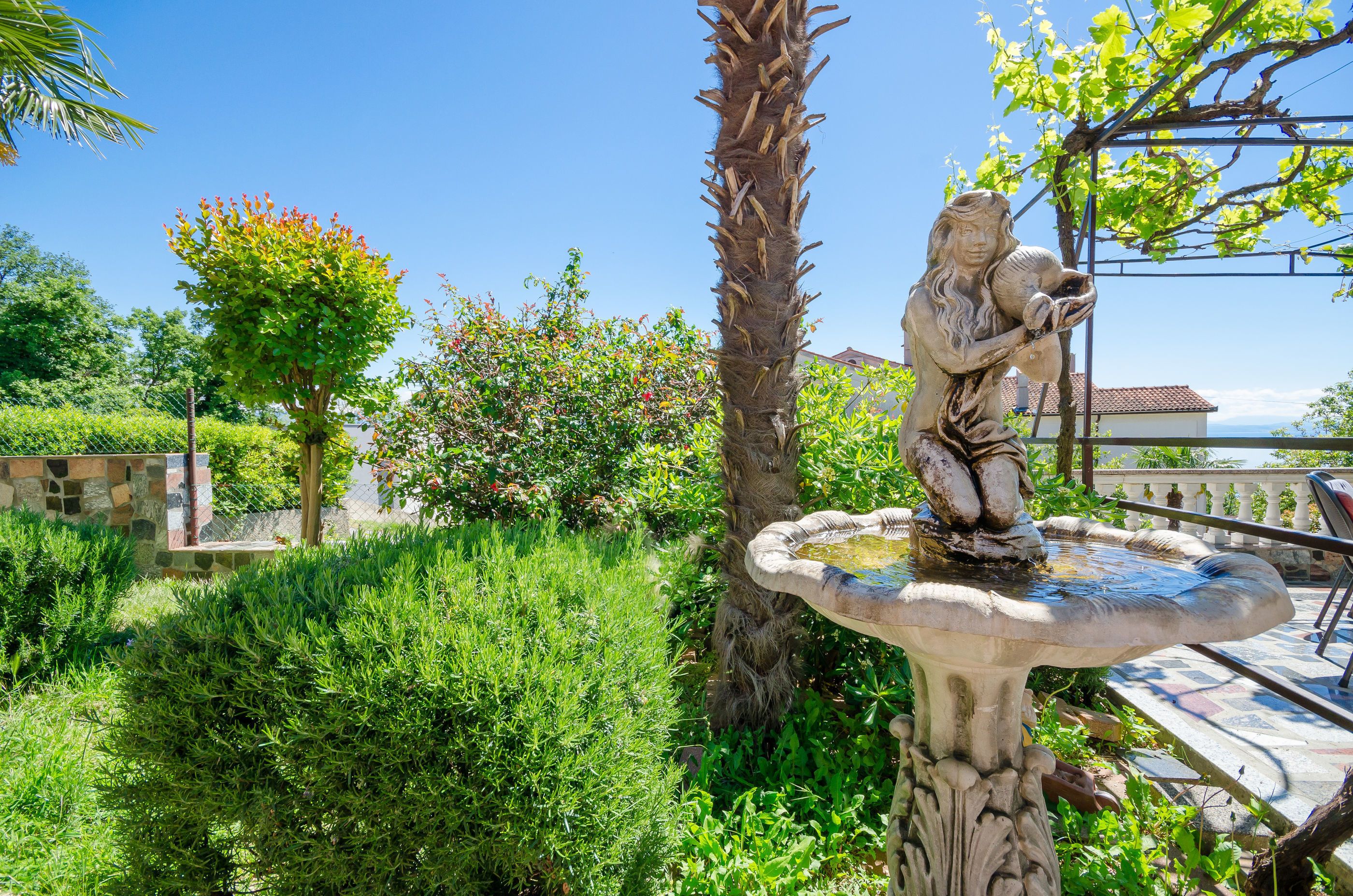Un jardin orné d’une statue représentant une femme tenant un oiseau, entouré d’une végétation luxuriante et surplombé d’un ciel bleu clair.