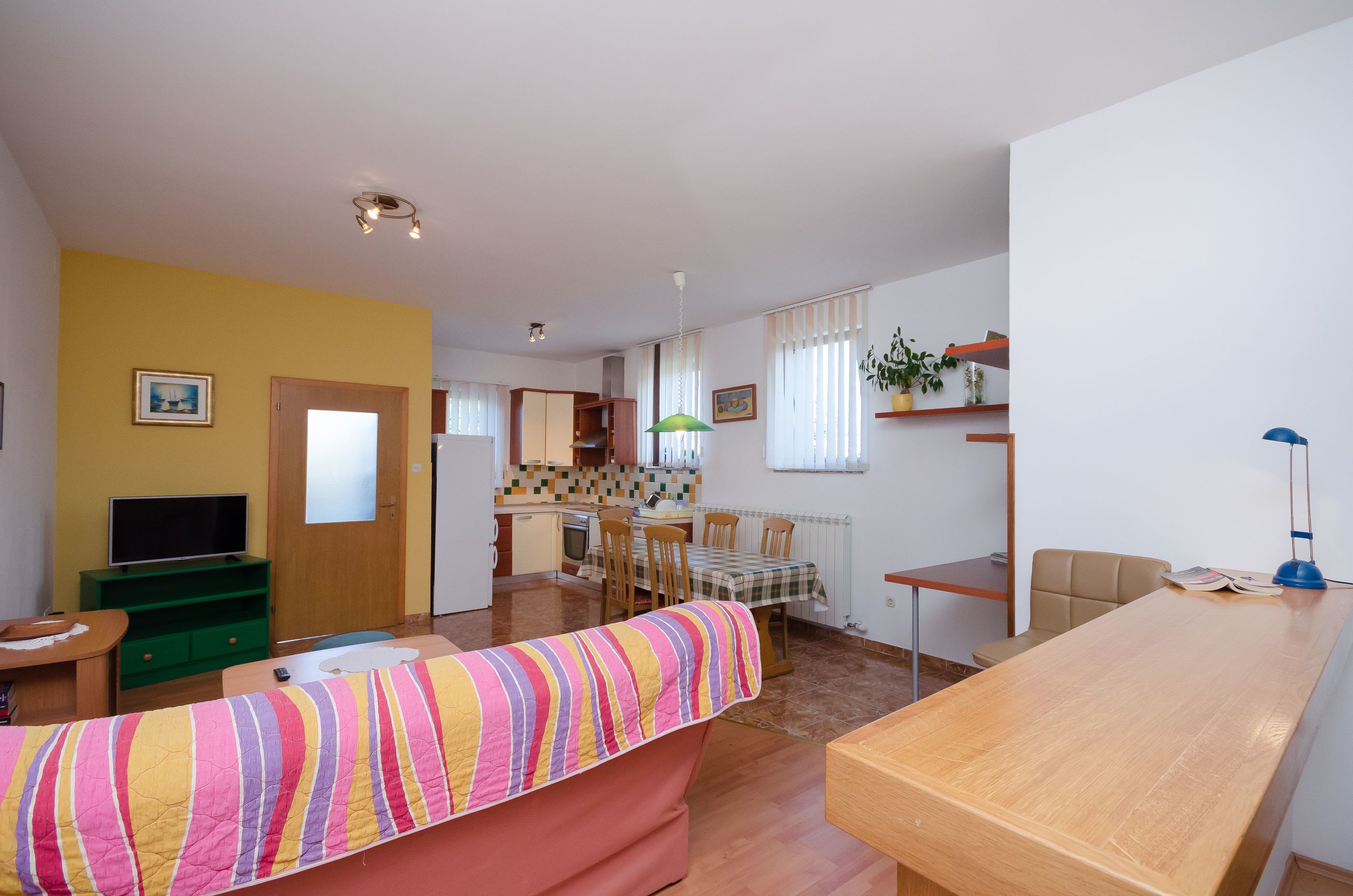 A living room with a sofa, a wooden table, a TV, and a kitchen in the background.